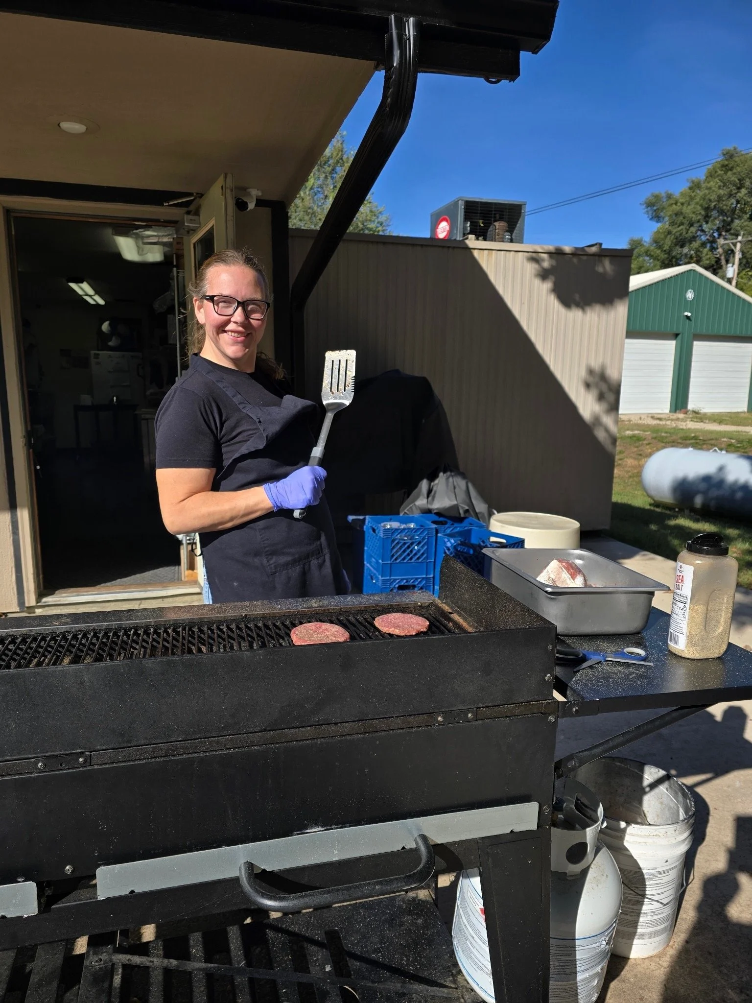 Elm Creek Beef Woman grilling hamburger patties on an outdoor grill, smiling at the camera, wearing glasses, a black shirt, and purple gloves.