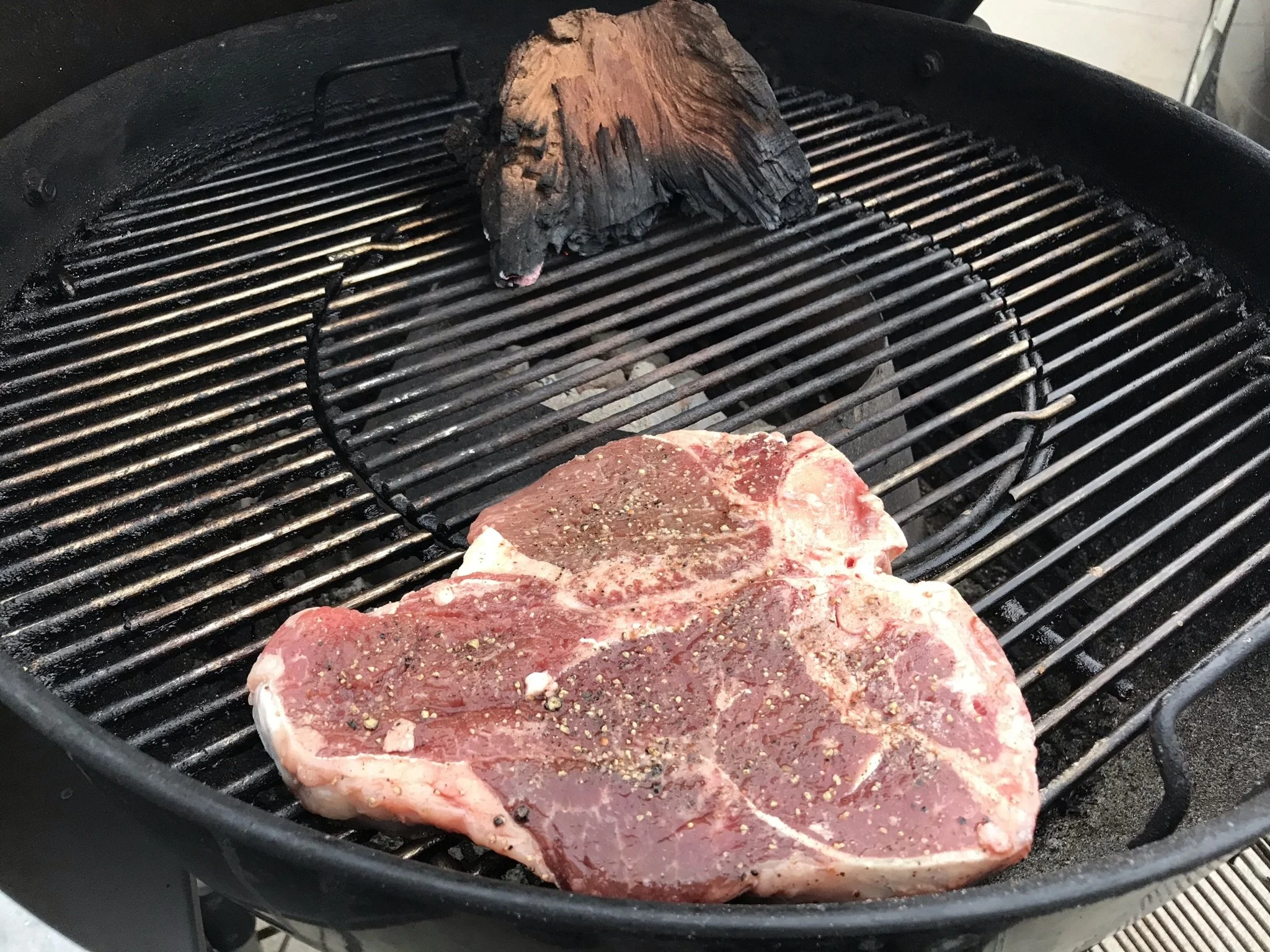 Elm Creek Beef Two raw steaks seasoned with salt and pepper cooking on a grill, with a piece of wood burning in the background.