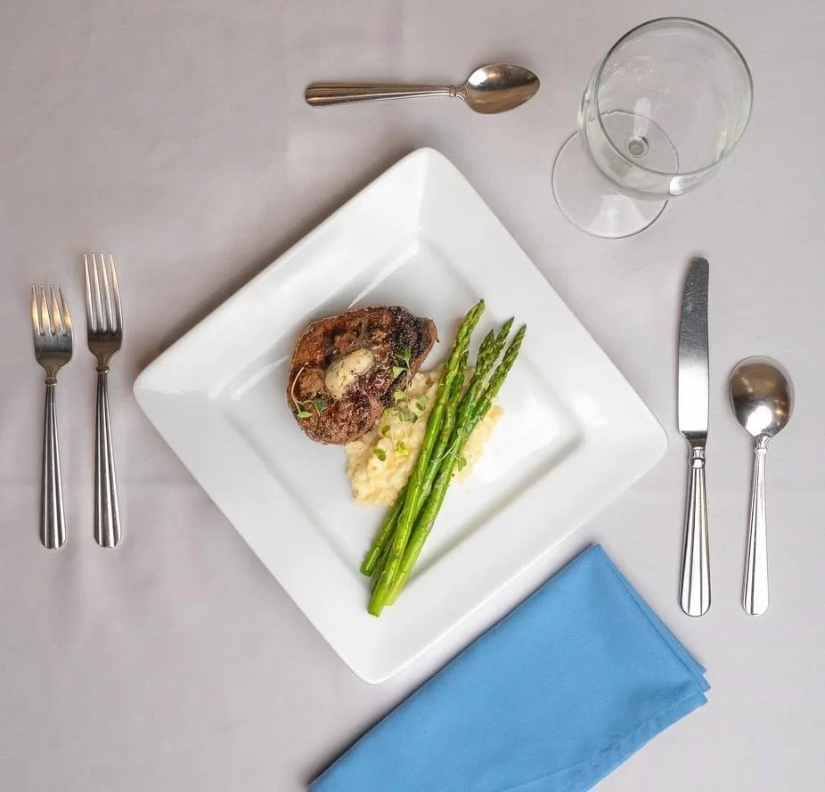 Elm Creek Beef A plate with a cooked steak, asparagus, and mashed potatoes, set on a table with utensils, a water glass, and a blue napkin.