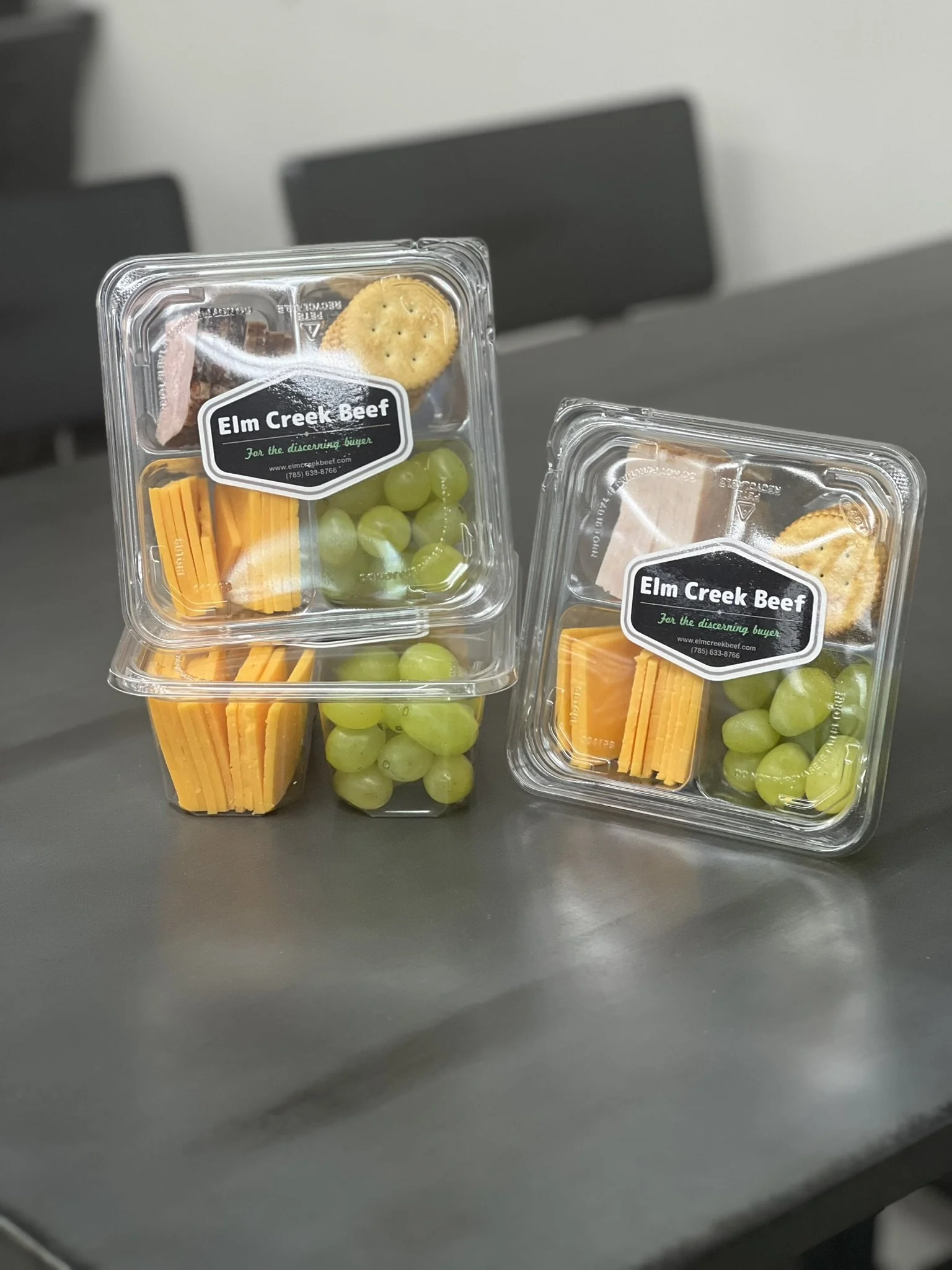 Elm Creek Beef Pre-packaged lunch containers with green grapes, cheddar cheese sticks, Ritz crackers, and beef slices, labeled 'Elm Creek Beef' on a dark table with black chairs in the background.