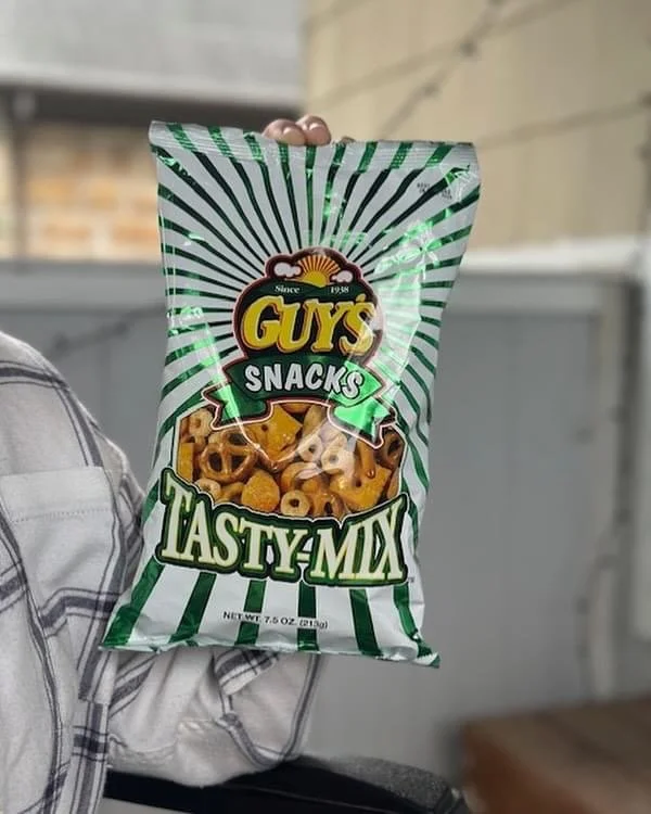 Elm Creek Beef A person holding a green and white striped bag of Guy's Snacks Tasty Mix, containing pretzels and cheese crackers, in an indoor setting.