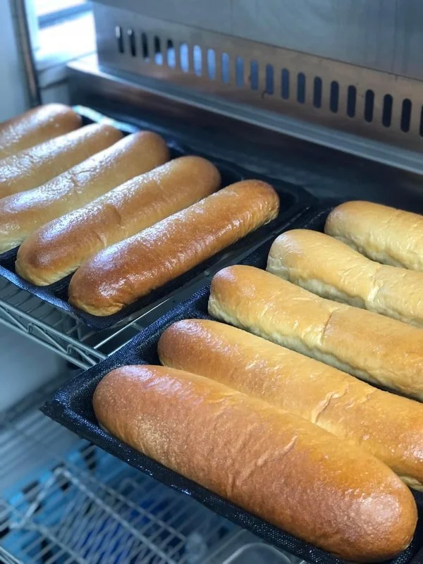 Close-up of several loaves of baked bread cooling on black trays inside a bakery oven. The loaves are elongated and golden brown.