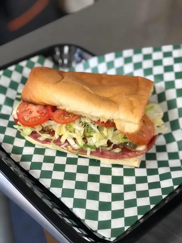 A sub sandwich with tomatoes, lettuce, and meat on a bun in a black basket lined with green and white checkered paper.