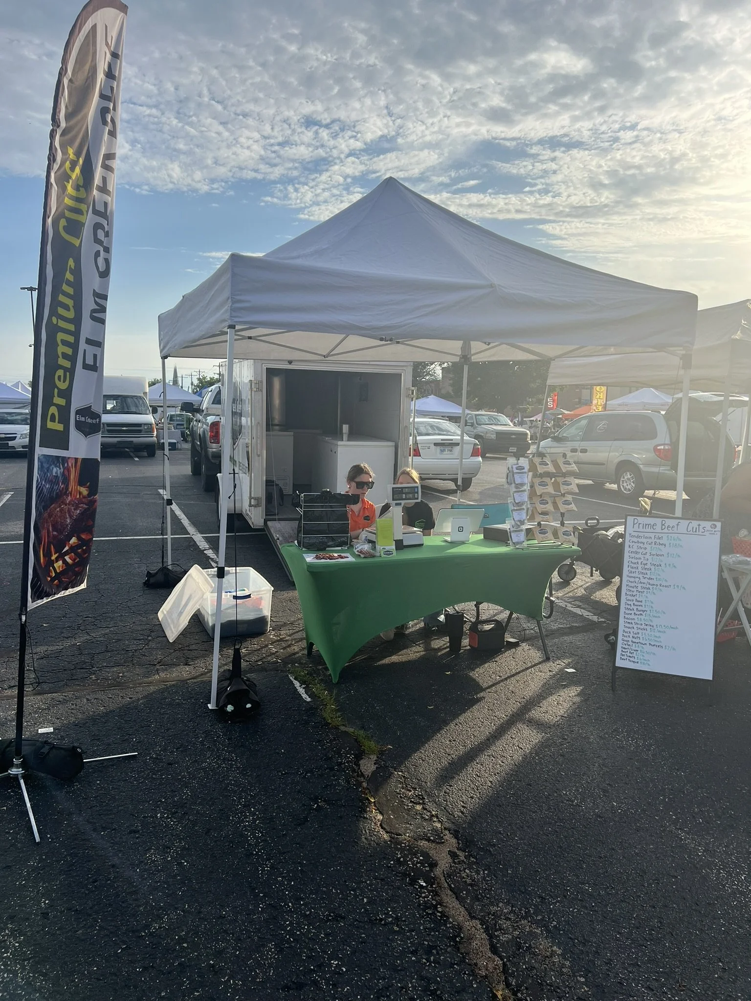 Elm Creek Beef A small outdoor market stall with a white canopy tent, green tablecloth, and a menu board, set up in a parking lot with cars and other tents in the background during sunset.