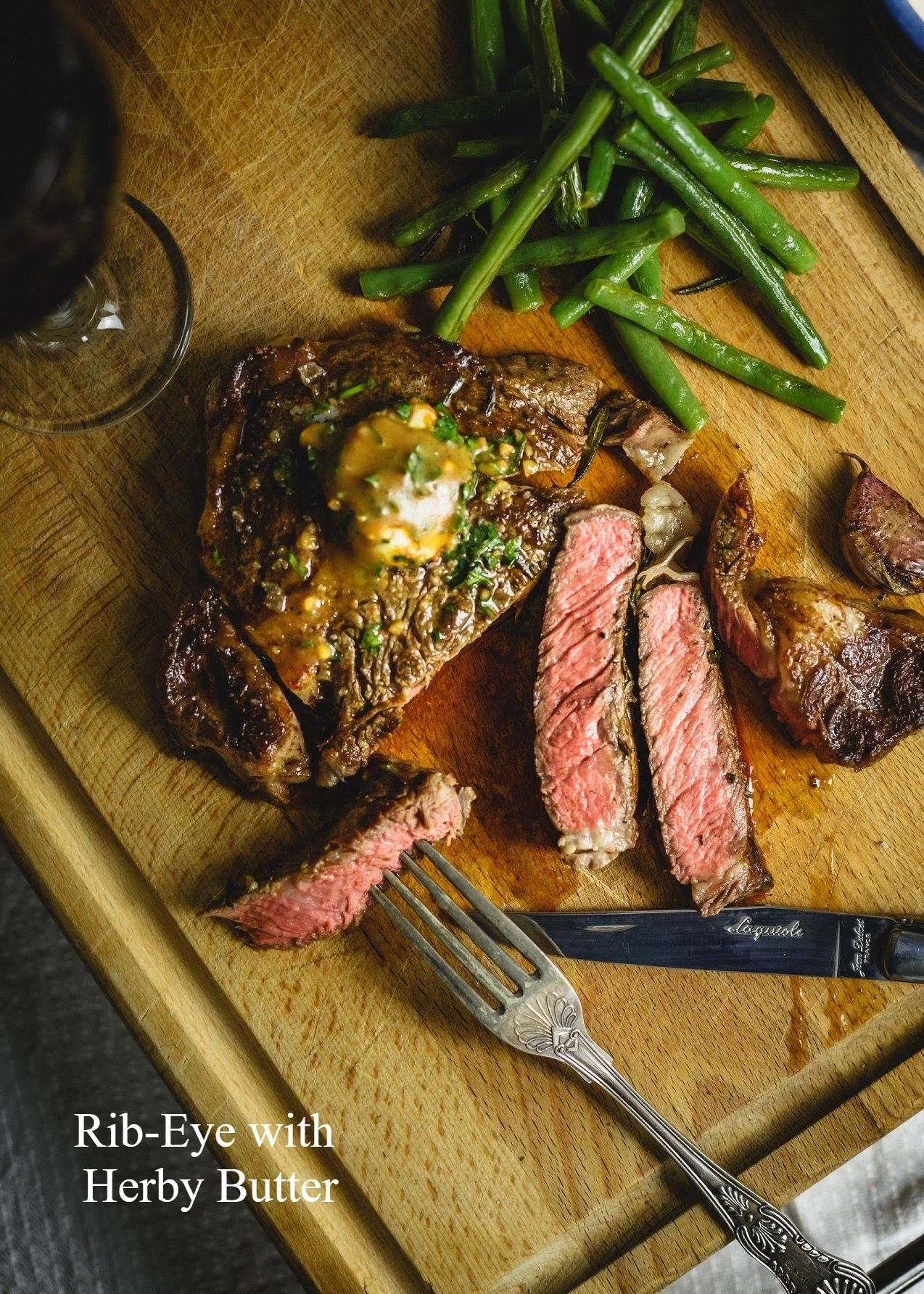 Elm Creek Beef A wooden cutting board with sliced cooked rib-eye steak topped with herb butter, green beans, a knife, and a fork.