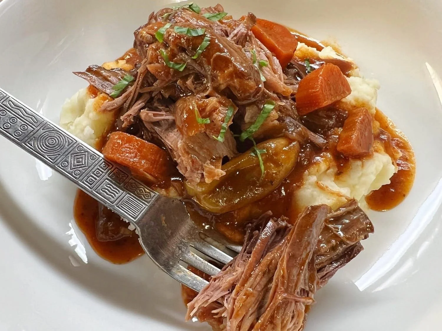 Elm Creek Beef A plate of mashed potatoes topped with beef stew containing shredded beef, carrots, and gravy, with a fork on the side.