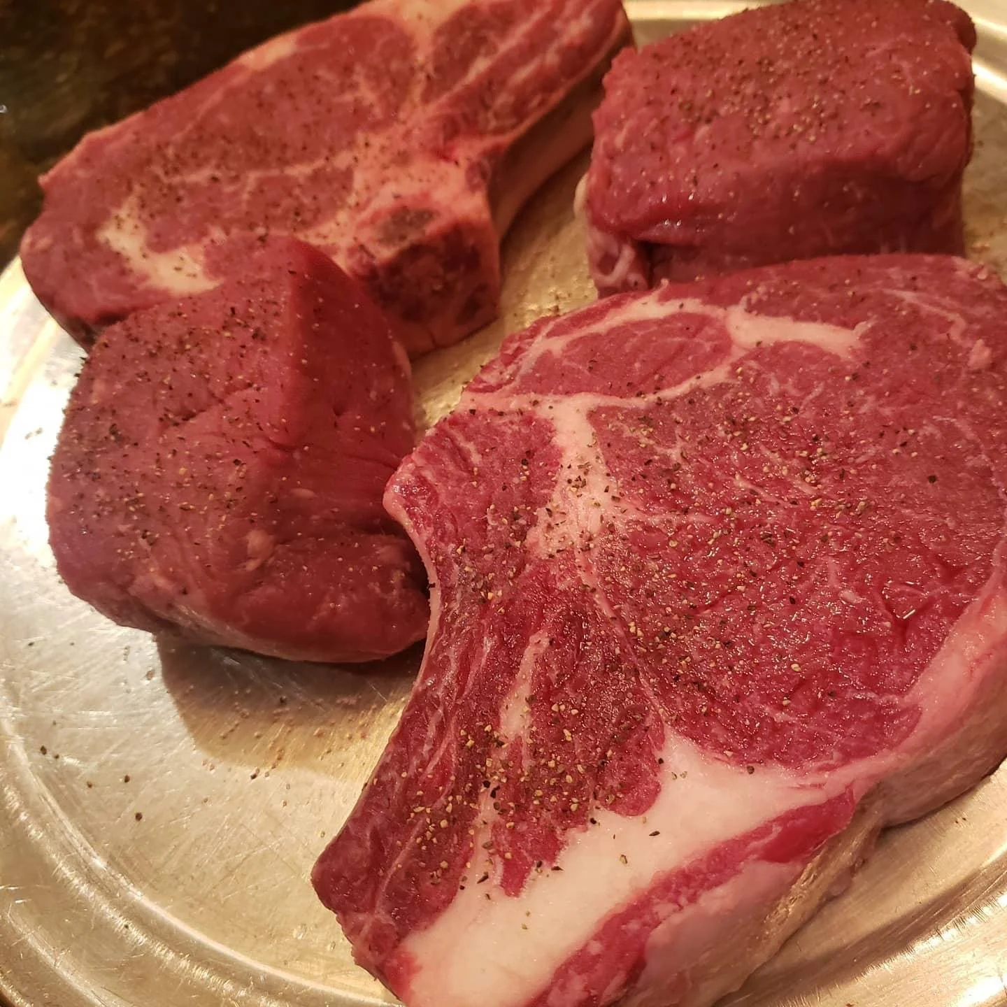 Elm Creek Beef Four raw beef steaks seasoned with salt and black pepper on a metal tray.