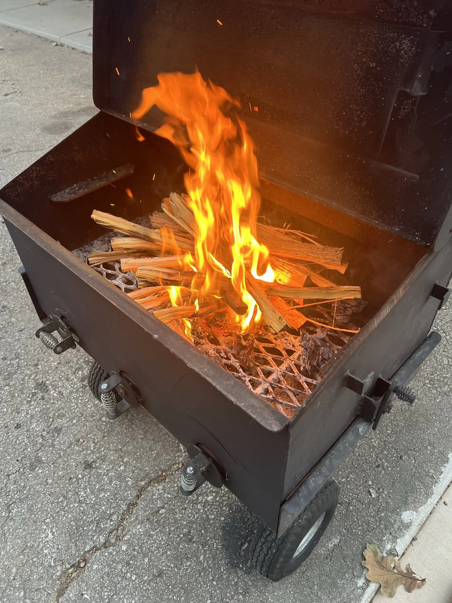 Elm Creek Beef A barbecue grill with an open lid, burning with bright flames and wood. The grill has wheels and is placed on a driveway.