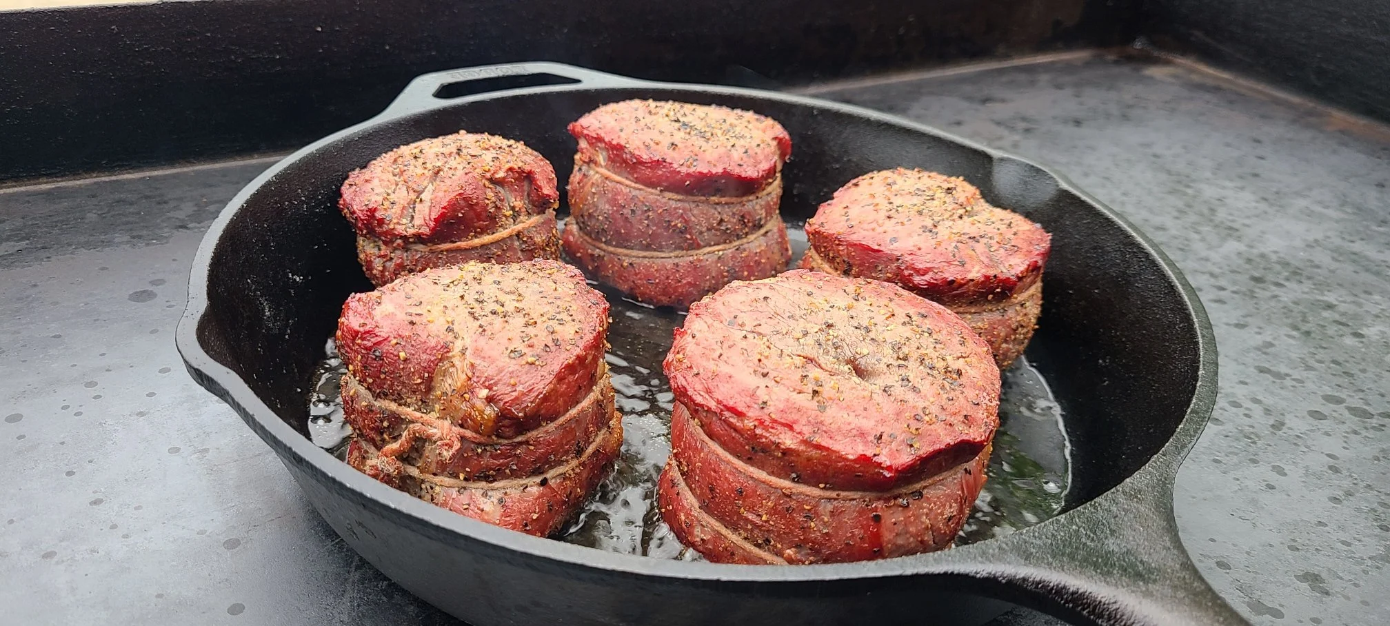 Elm Creek Beef Six thick cuts of raw meat seasoned with herbs and black pepper, tied with kitchen twine, cooking in a cast iron skillet on a stovetop.
