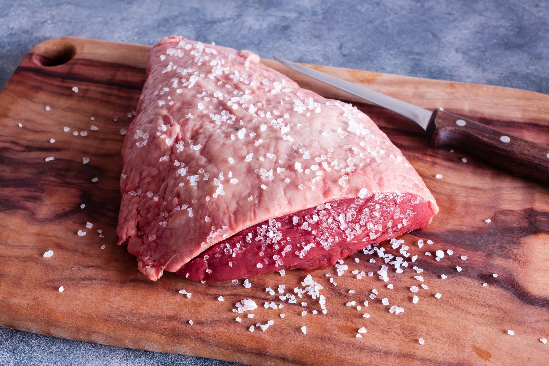 Elm Creek Beef A raw cut of beef on a wooden cutting board, sprinkled with coarse salt, with a kitchen knife nearby.