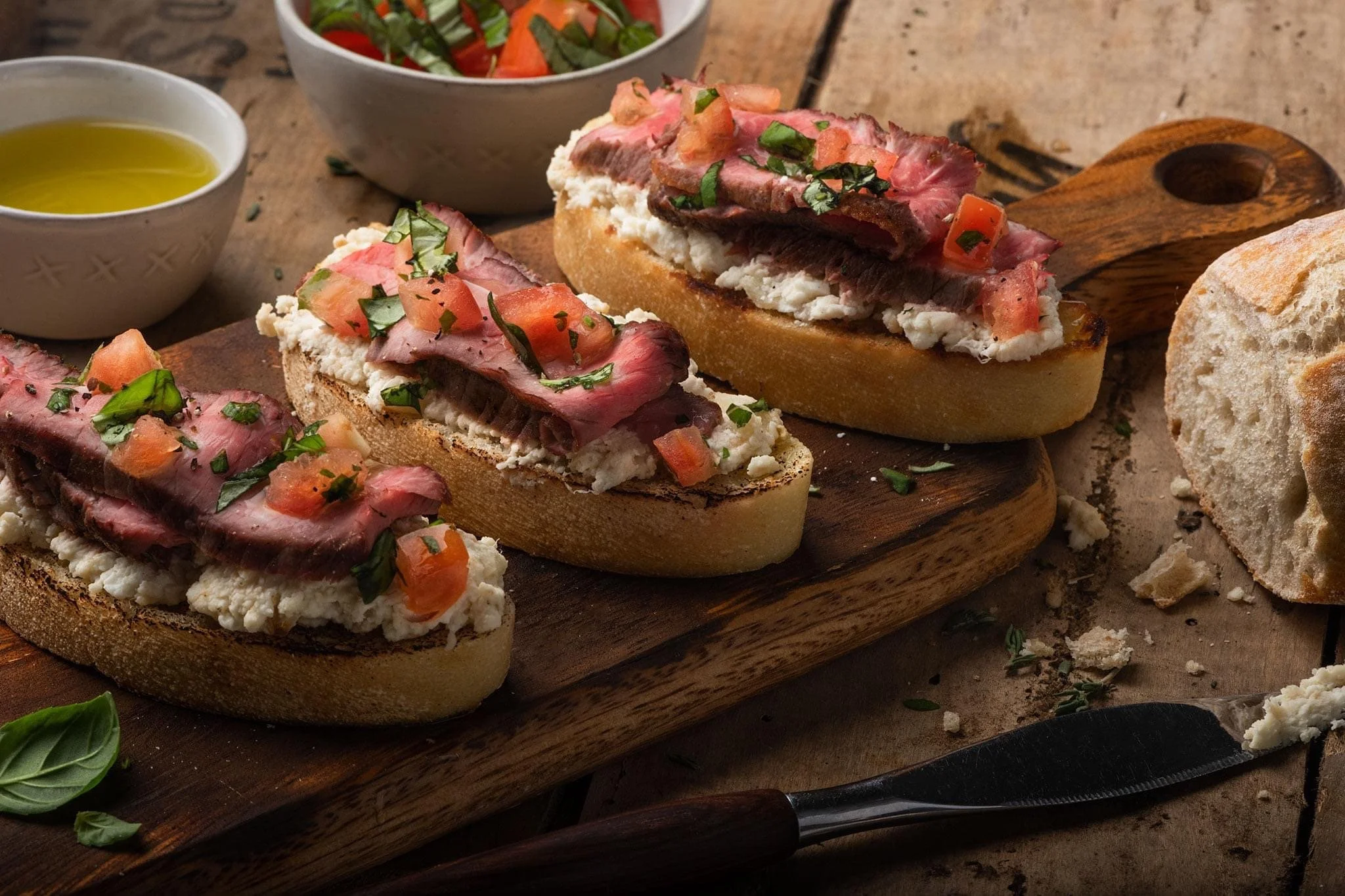 Elm Creek Beef Open-faced sandwiches on a wooden cutting board with bread, cheese, slices of cooked beef, chopped tomatoes, basil, and herbs, with a side salad, a knife, and small bowls of olive oil and vinegar.