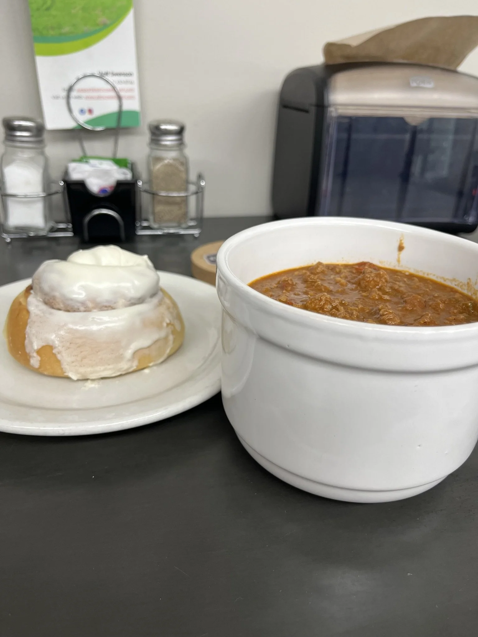 Elm Creek Beef A bowl of chili and a cinnamon roll with icing on a dark table in a restaurant or cafe setting.