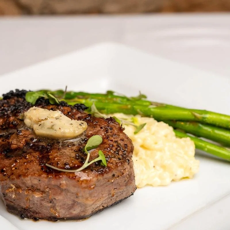 Elm Creek Beef A plated meal with a grilled steak topped with garlic, mashed potatoes, and cooked green asparagus.