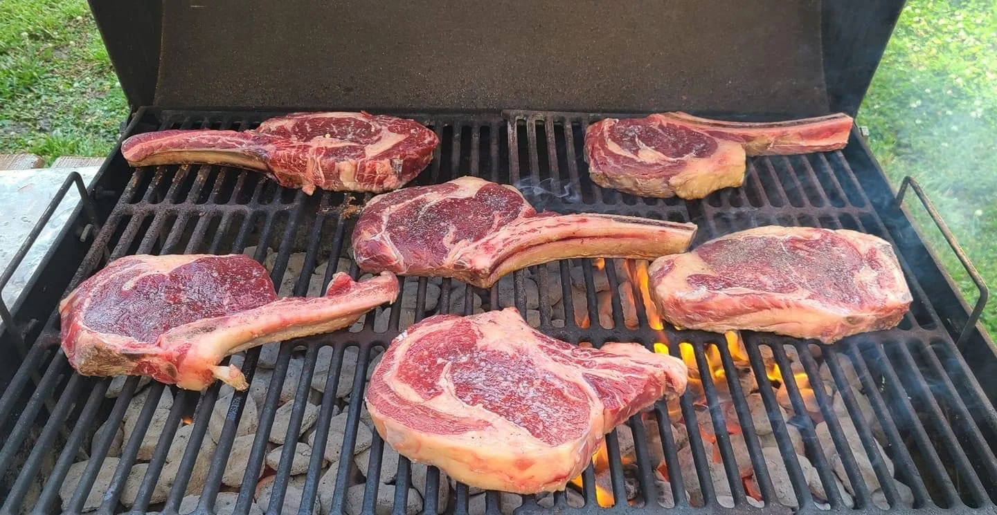 Elm Creek Beef Six raw ribeye steaks cooking on a charcoal grill, with some flames and smoke visible.