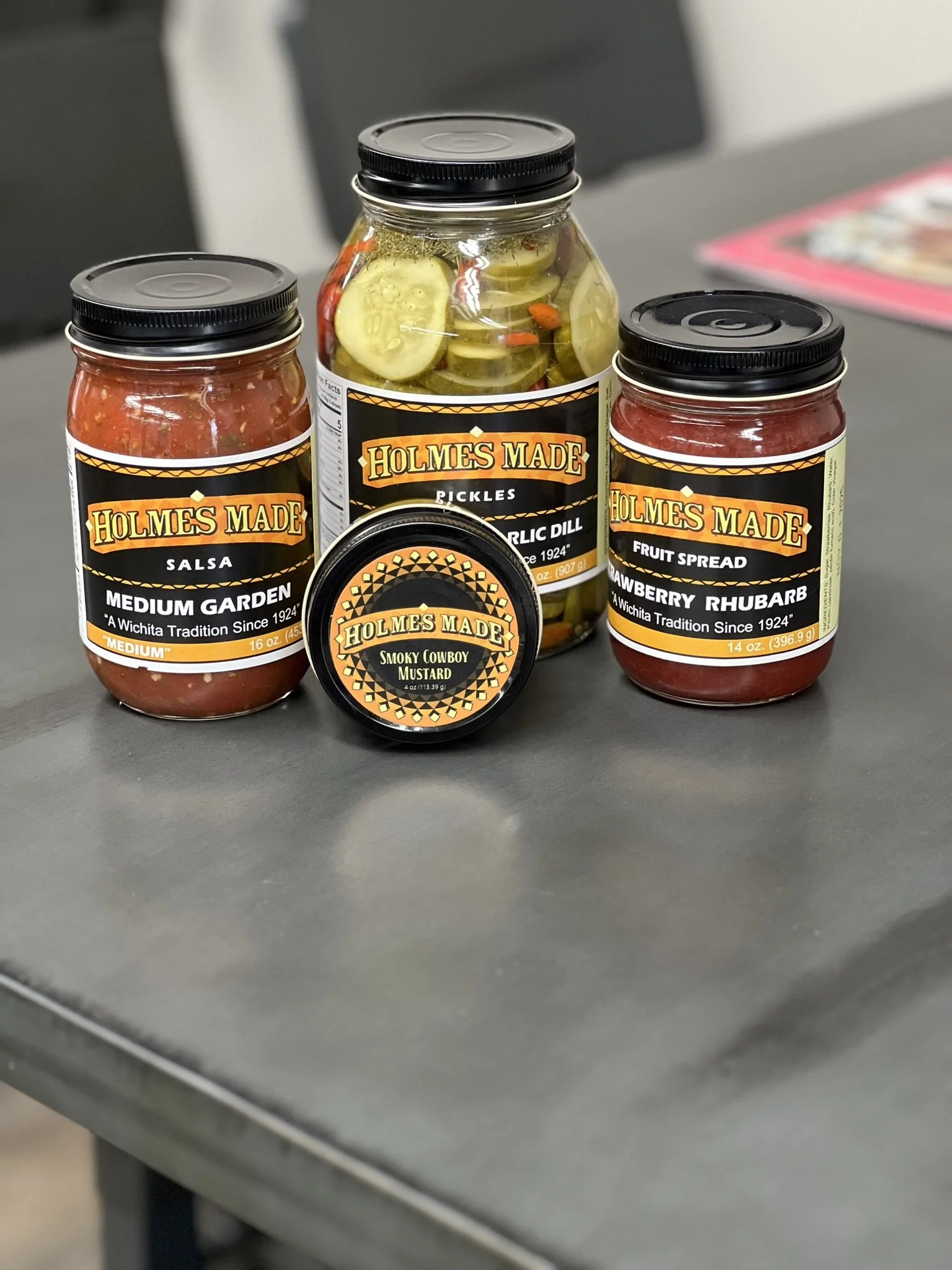 Elm Creek Beef Collection of five jars and a small container of Holmes Made brand food items on a gray table, including salsa, pickles, fruit spread, smokey cowboy mustard, and a jar of mustard.