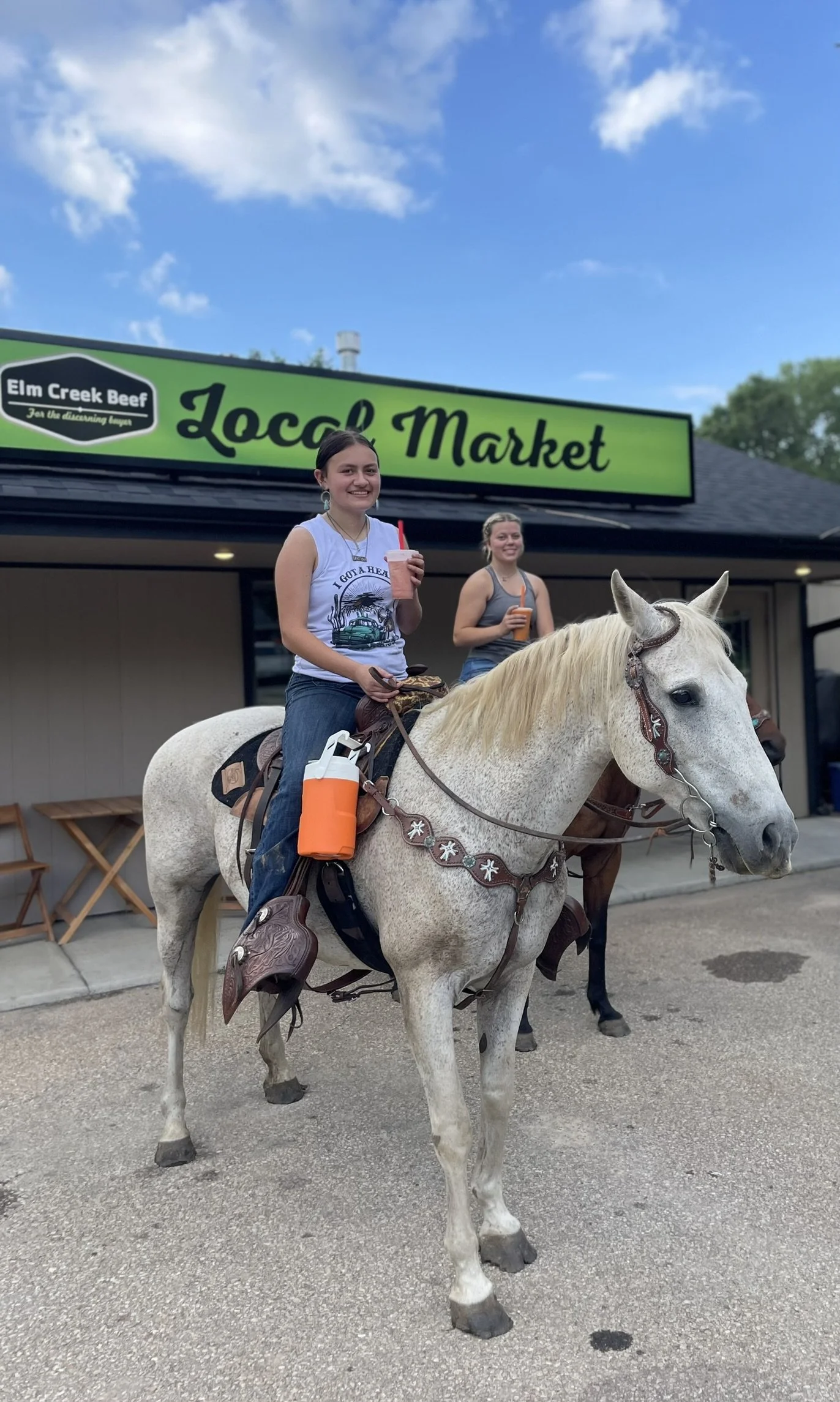 Elm Creek Beef Two women riding a white horse in front of a green storefront with a sign saying 'Local Market' and 'Elm Creek Beef'. One woman is smiling, holding a drink, and the other is in the background also holding a drink.
