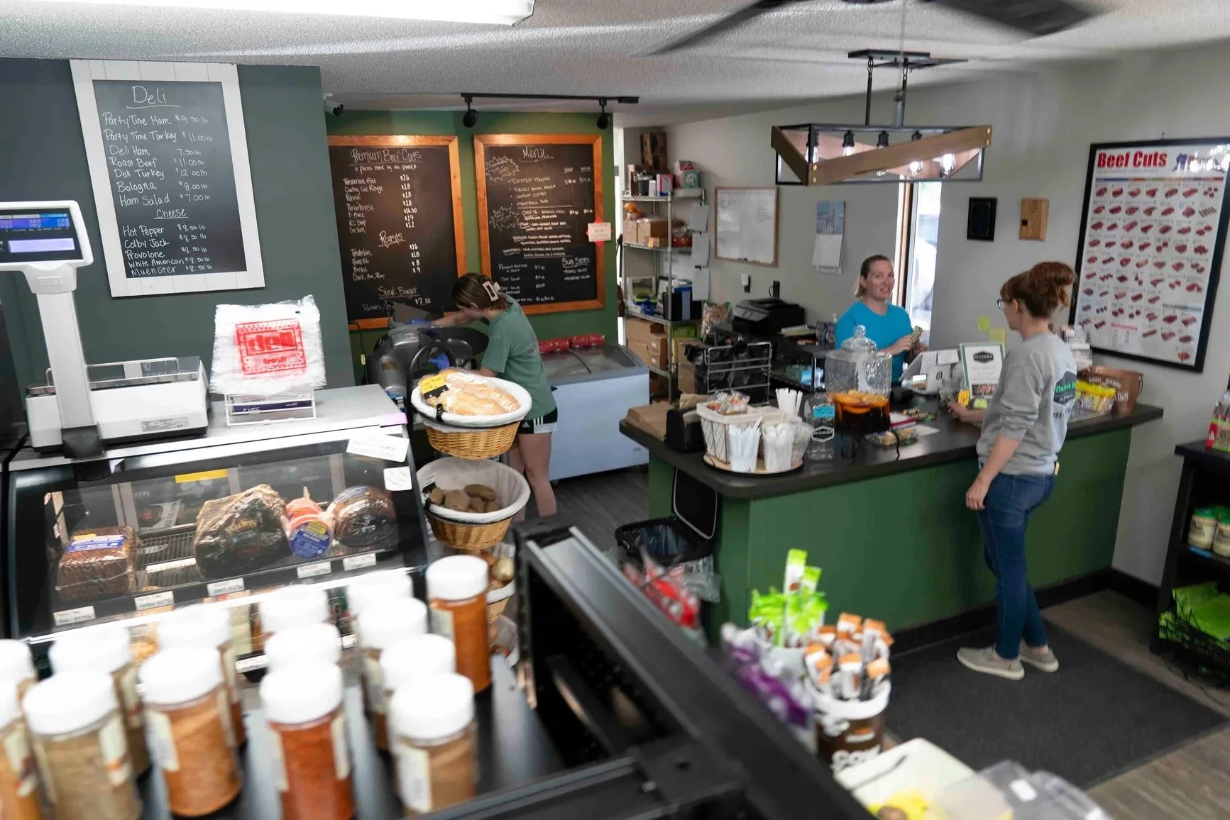 Elm Creek Beef Interior of a small deli or sandwich shop with a green and white counter, menu boards on the wall, a cash register, a scale, and staff members preparing food and taking orders.