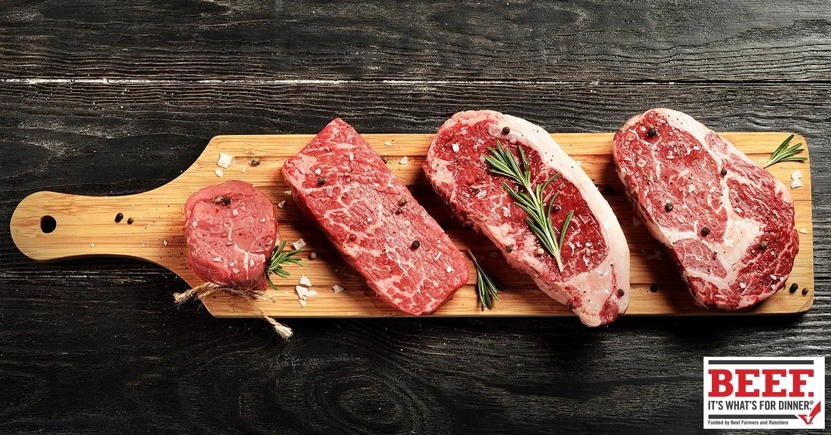 Elm Creek Beef Four raw steaks with marbling on a wooden cutting board, garnished with rosemary sprigs, placed on a dark wooden surface.