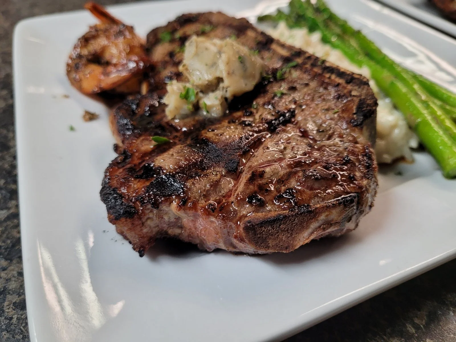 Elm Creek Beef Grilled steak with char marks, topped with a dollop of herb butter, served with grilled green asparagus and mashed potatoes on a white square plate.