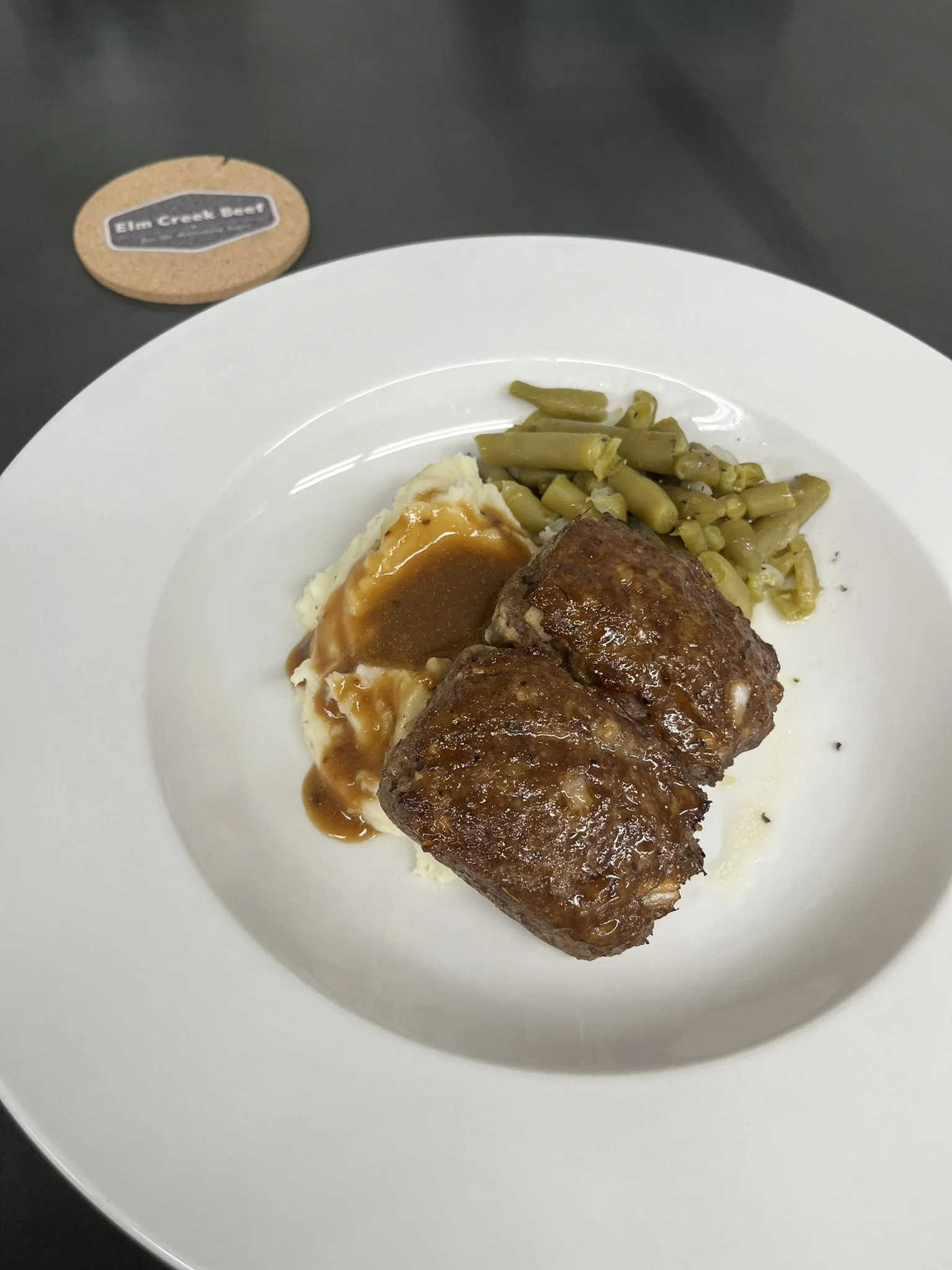 Elm Creek Beef Plate of mashed potatoes with gravy, green beans, and two pieces of cooked meatloaf. In the background, a coaster with a label that reads 'Elm Creek Beer'.