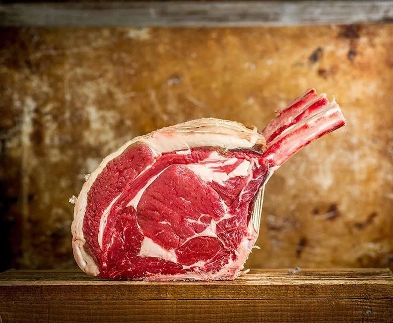 Elm Creek Beef A raw beef prime rib roast with a large bone, placed on a wooden surface against a rustic background.