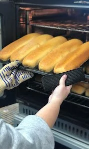 Person wearing an oven mitt placing a tray of hot dog buns into an oven.
