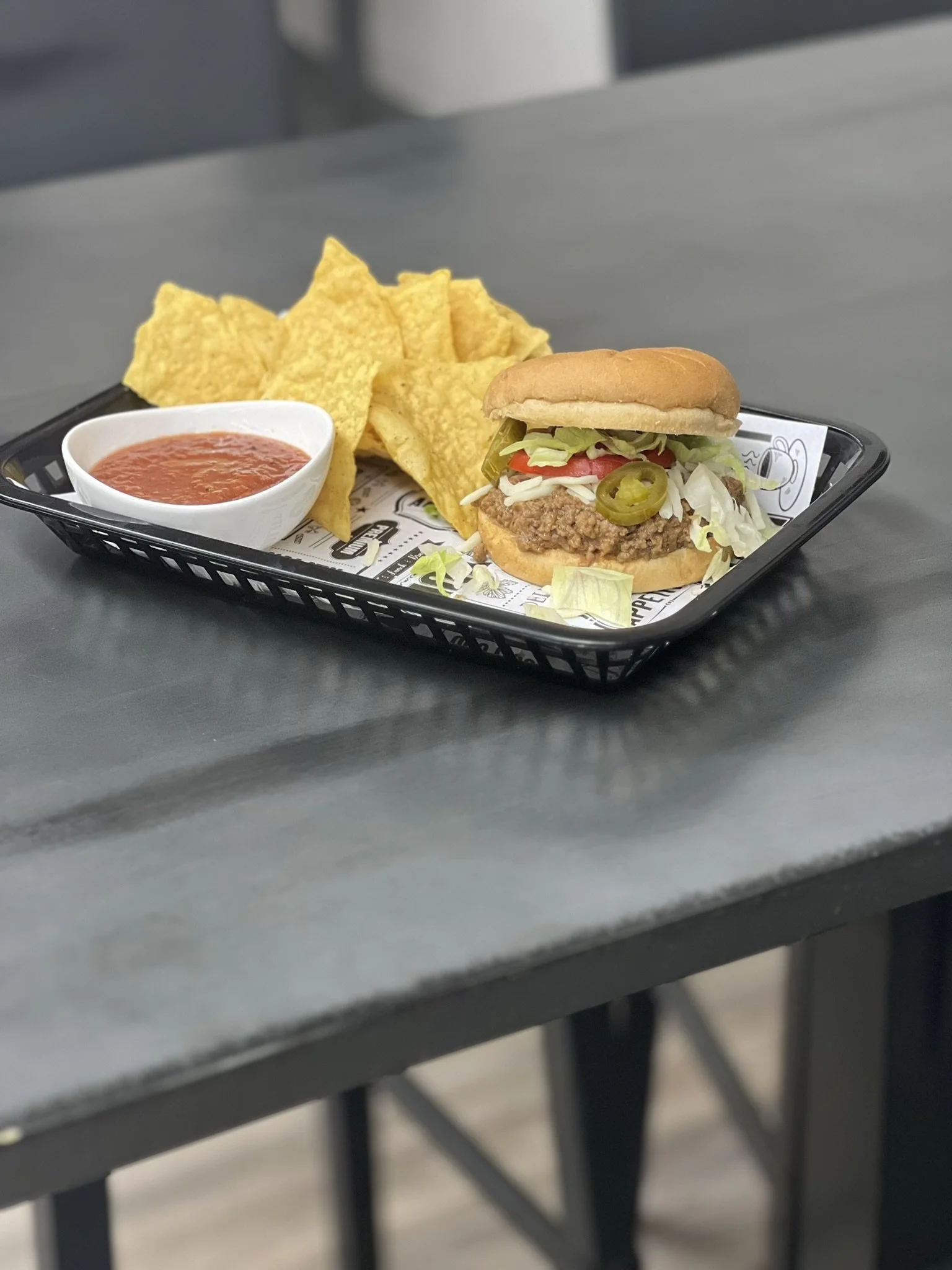 Elm Creek Beef A black tray with a hamburger, tortilla chips, and a bowl of salsa on a gray table.