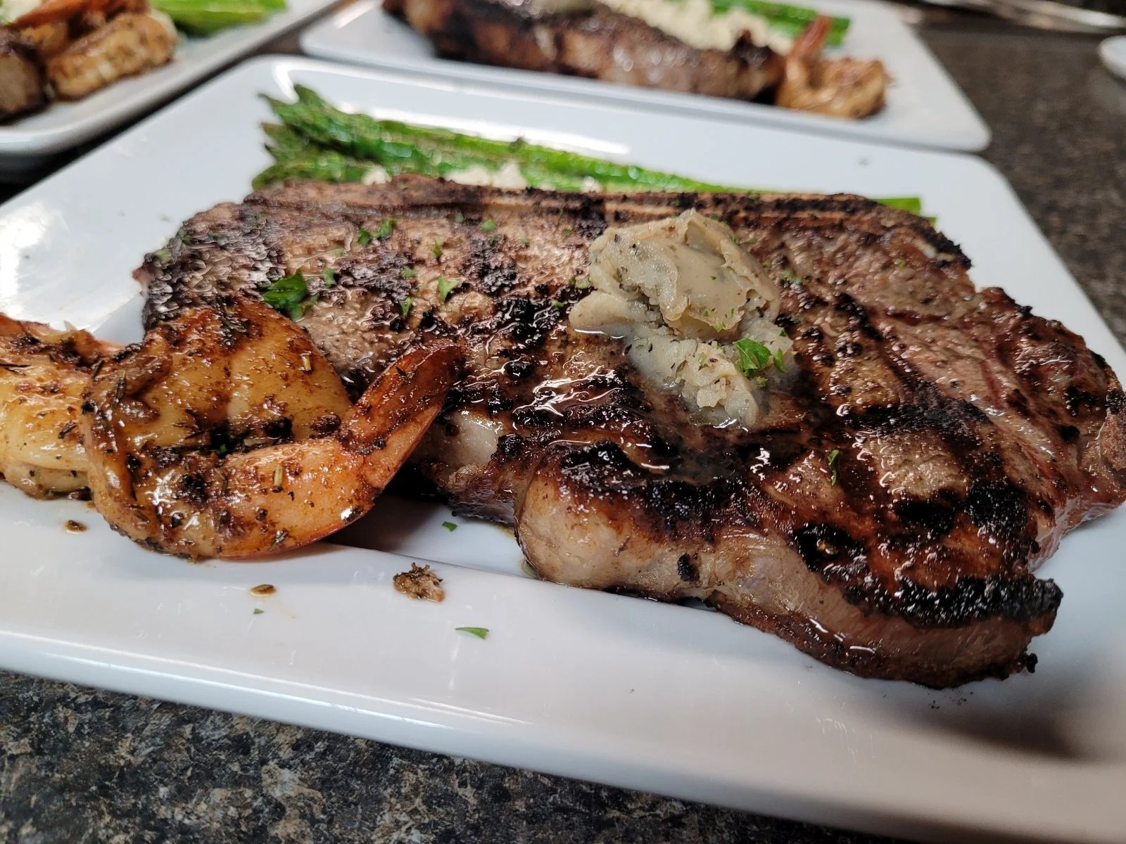 Elm Creek Beef Grilled steak with a dollop of butter, grilled shrimp, green asparagus, and roasted potatoes on a white plate.