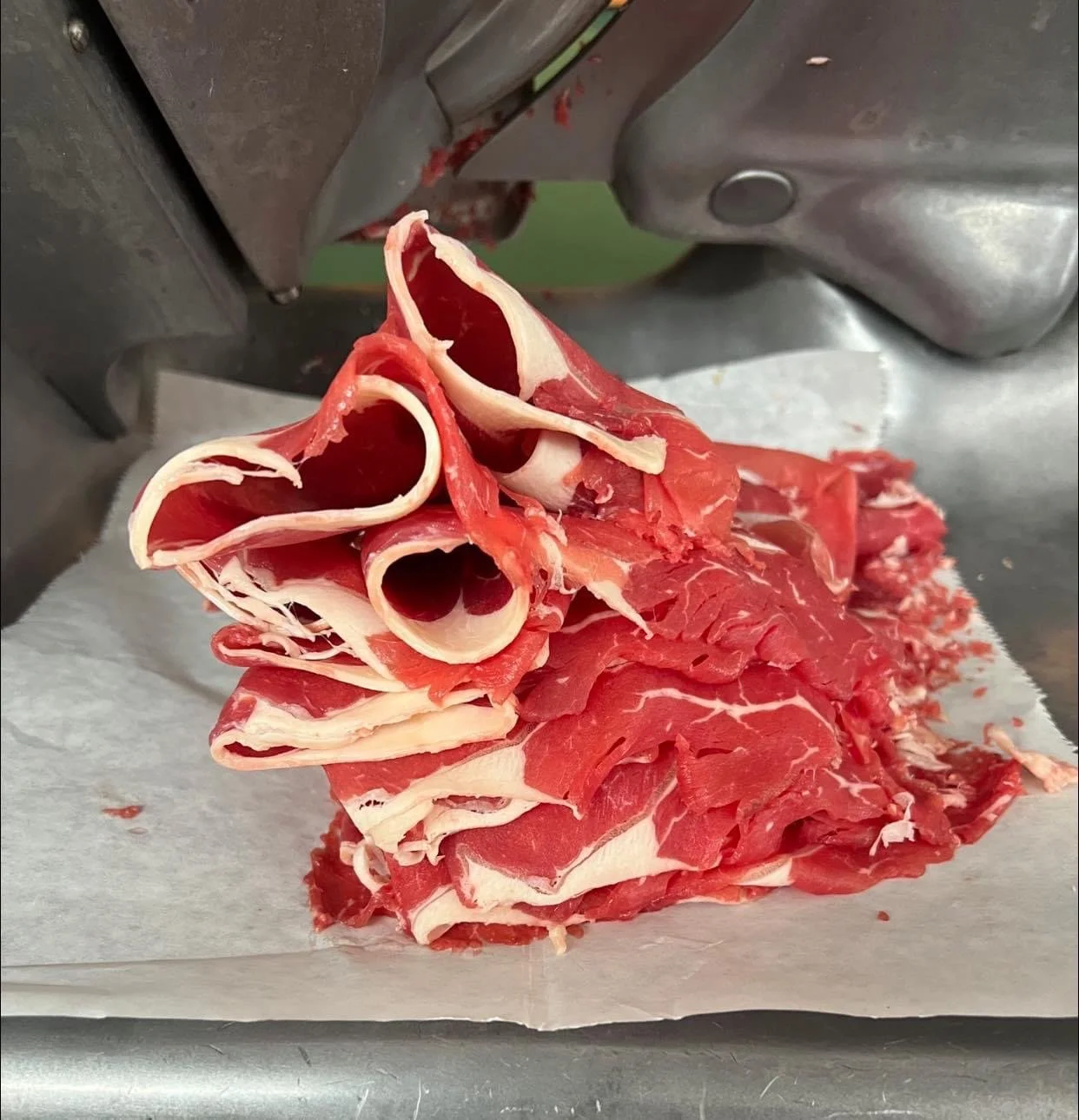 Elm Creek Beef Thin slices of raw beef arranged in a pile on white parchment paper inside a meat slicer.
