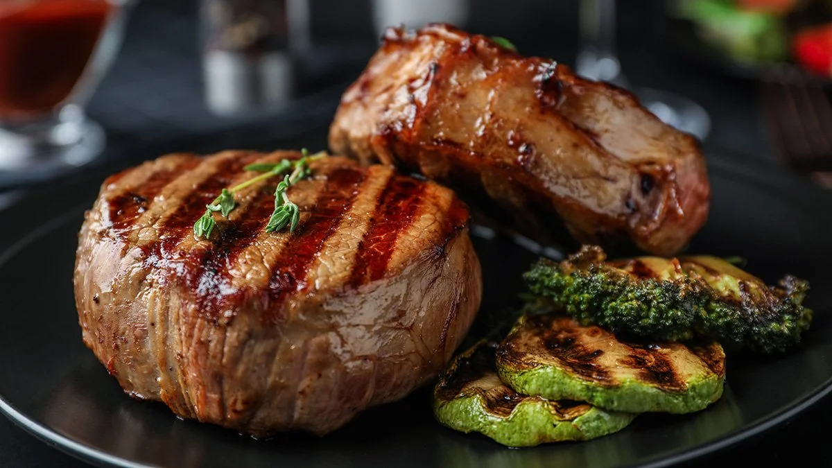 Elm Creek Beef Grilled steak topped with herbs, accompanied by grilled vegetables, including zucchini, broccoli, and bell pepper, on a black plate.