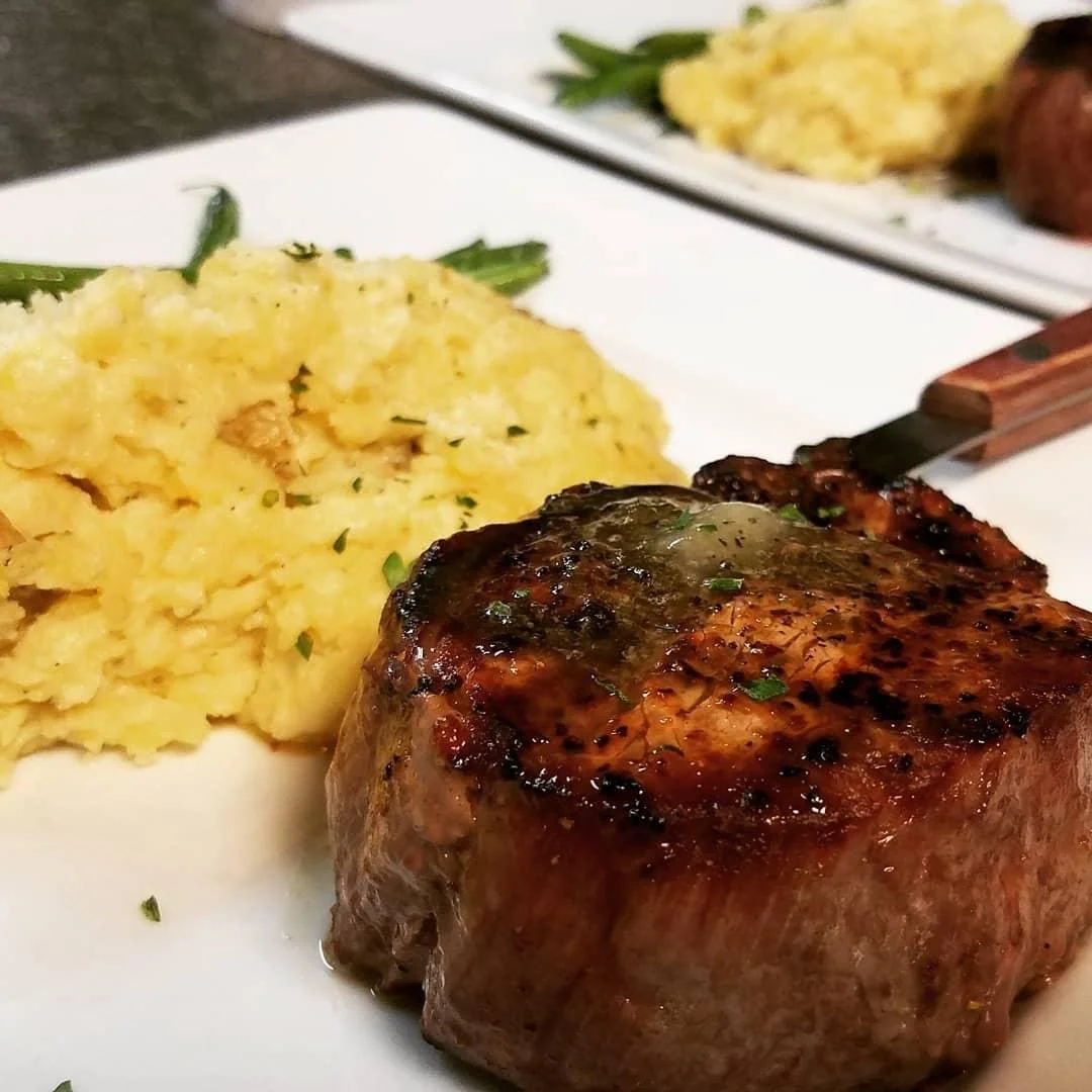 Elm Creek Beef Close-up of a plated meal with mashed potatoes garnished with herbs, and a grilled steak with a bone, seasoned and cooked to a brown color, with another plate of mashed potatoes in the background.