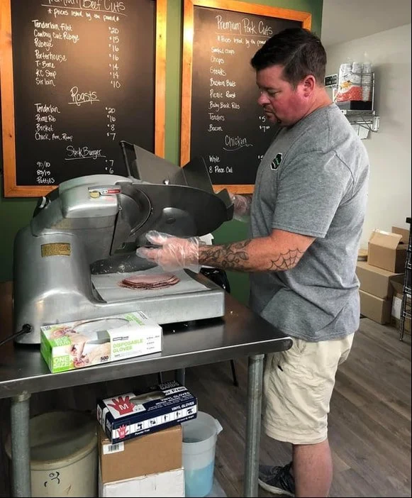 A man with tattoos on his left arm wearing a gray T-shirt and khaki shorts slices deli meat using a meat slicer at a counter in a grocery store or deli. There are blackboards with menu items on the wall behind him.