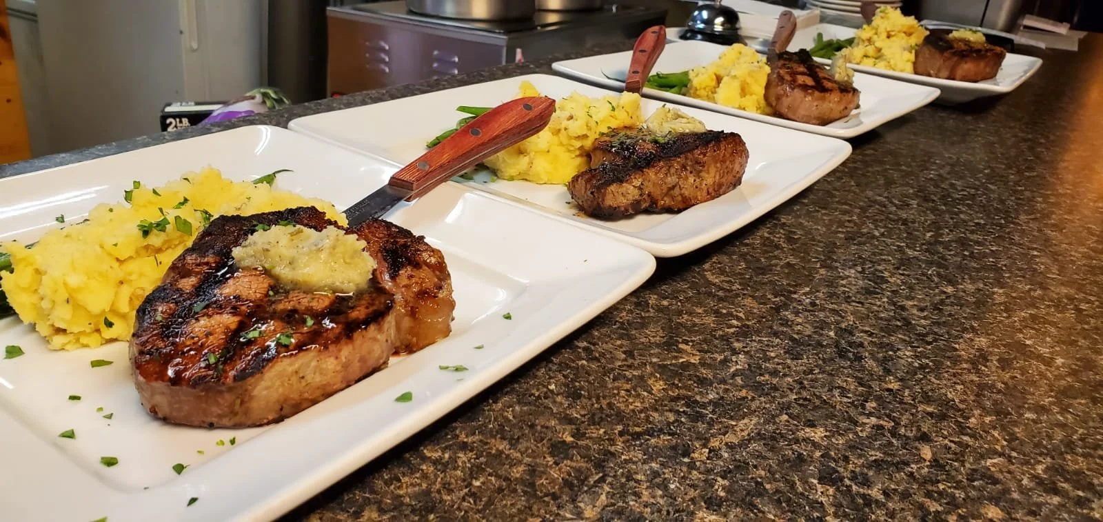 Elm Creek Beef Four white plates with grilled pork chops, mashed potatoes, and green beans on a dark granite countertop, arranged in a row.