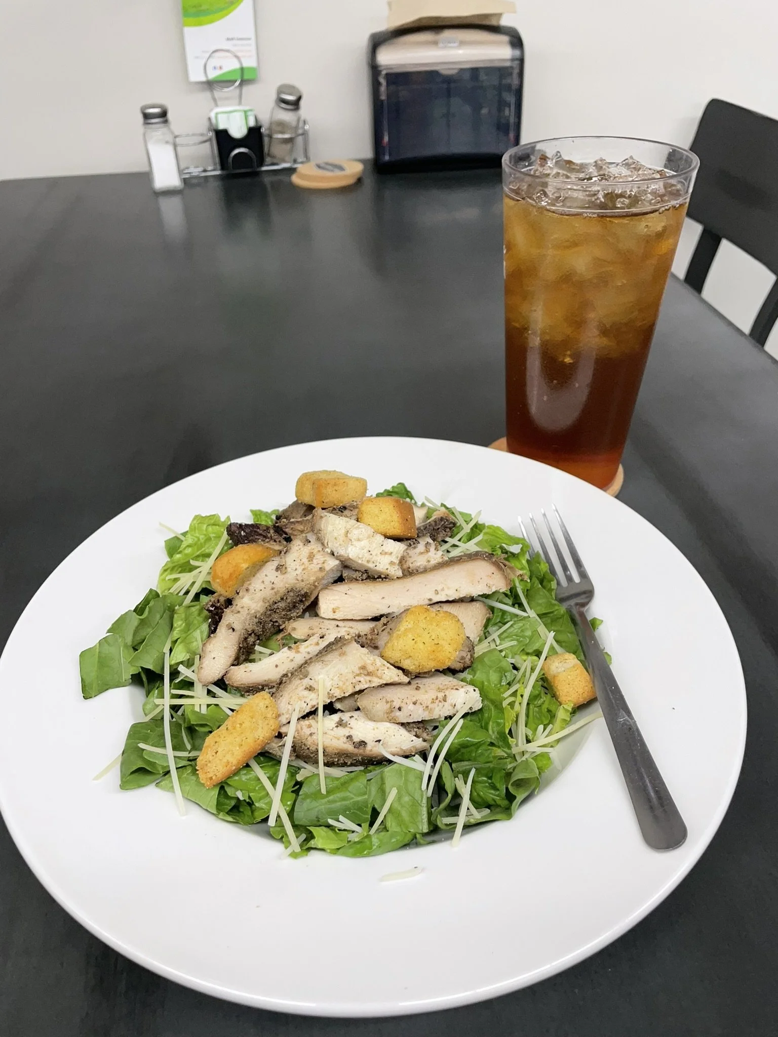Elm Creek Beef A salad with sliced grilled chicken, croutons, and shredded cheese on a bed of lettuce, with a tall glass of iced tea on a black table.