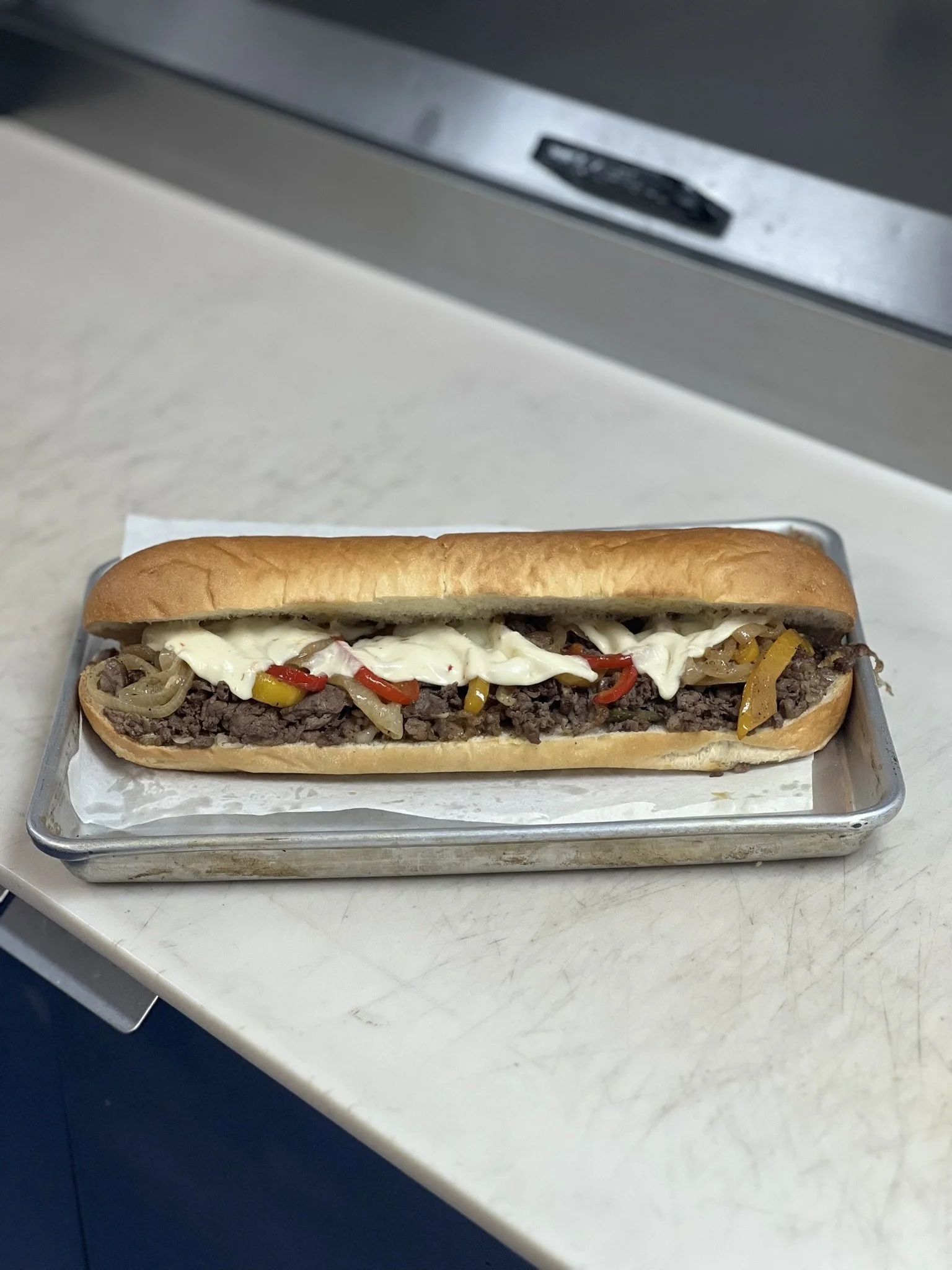 Elm Creek Beef A hot dog with chopped onions, yellow and red peppers, mayonnaise or mustard, in a soft bun on a metal tray.