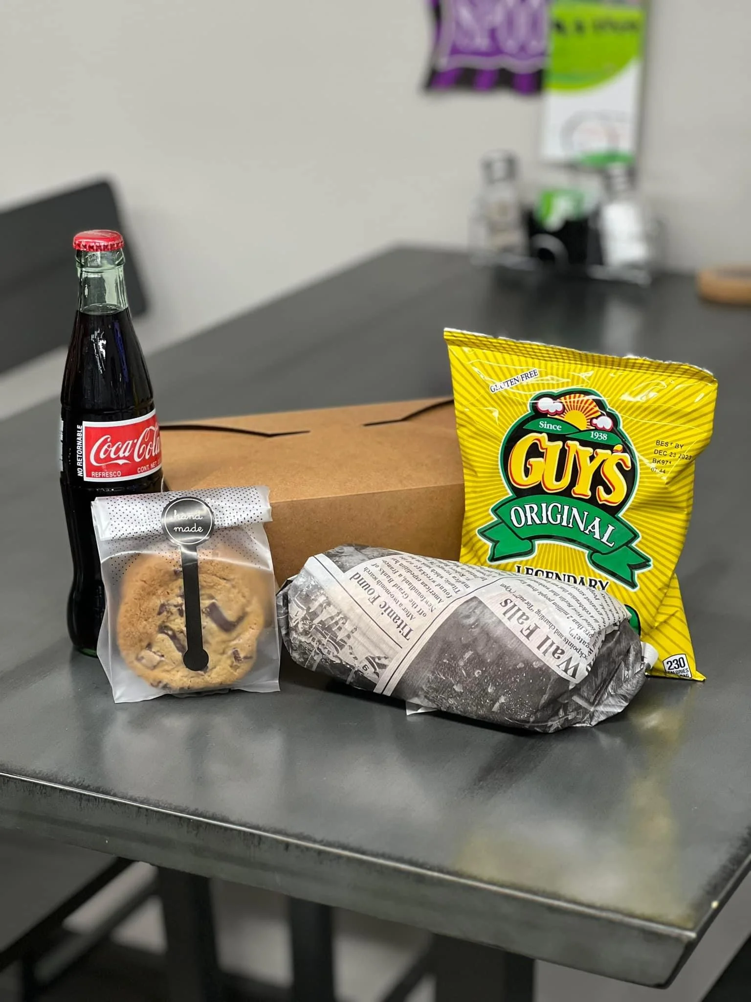 Elm Creek Beef A Coca-Cola glass bottle, a bag of cookies, a wrapped sandwich, and a bag of Goya brand potato chips on a gray table.