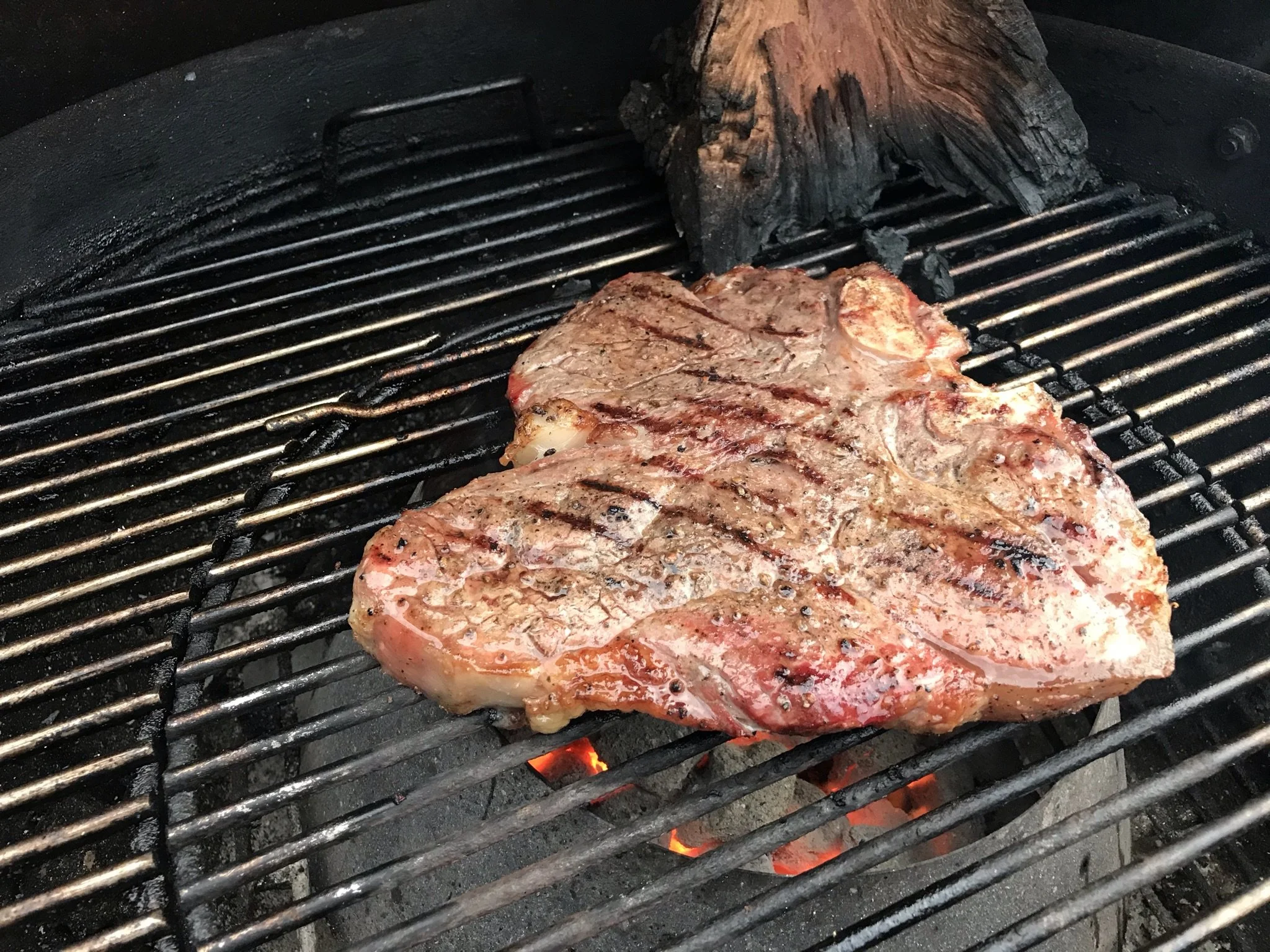 Elm Creek Beef A thick cut of steak cooking on a charcoal grill with visible grill lines, small flames, and a piece of wood in the background.