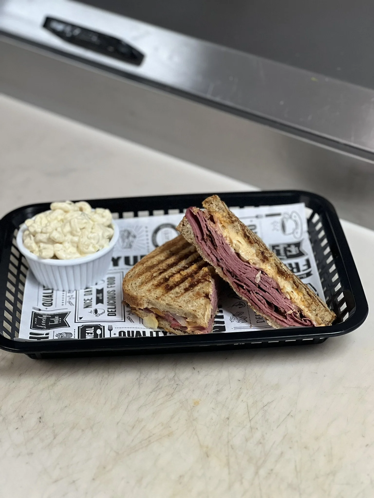 Elm Creek Beef Grilled sandwich with sliced corned beef and melted cheese, served with a side of macaroni salad in a small white bowl on a black tray lined with paper featuring black and white printed text and graphics.