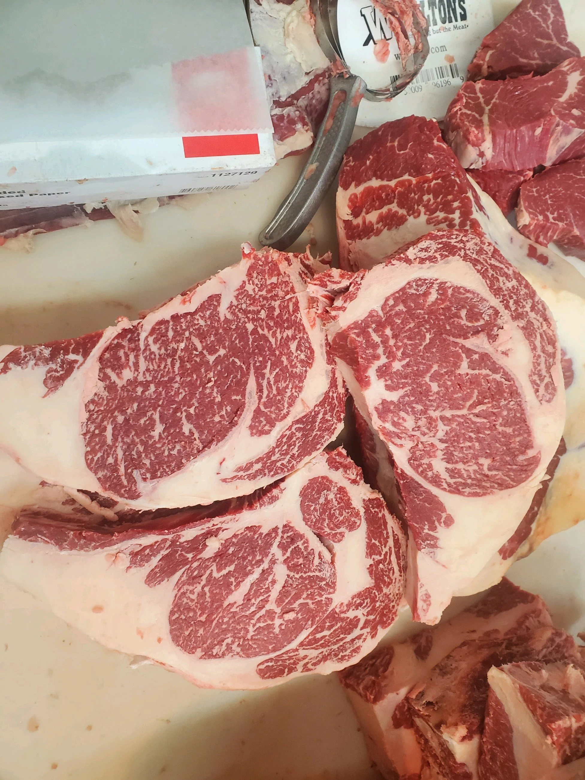 Elm Creek Beef Several cuts of raw marbled beef, likely steaks, on a white surface with a meat cutter and packaging in the background.