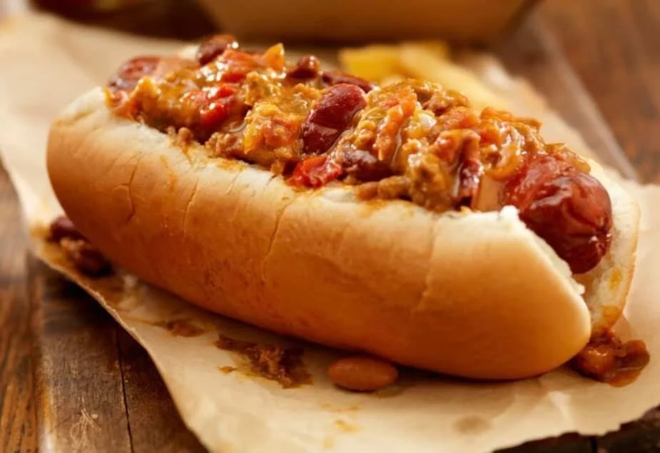 Elm Creek Beef Hot dog in a bun with chili, cheese, and beans on a wooden surface.