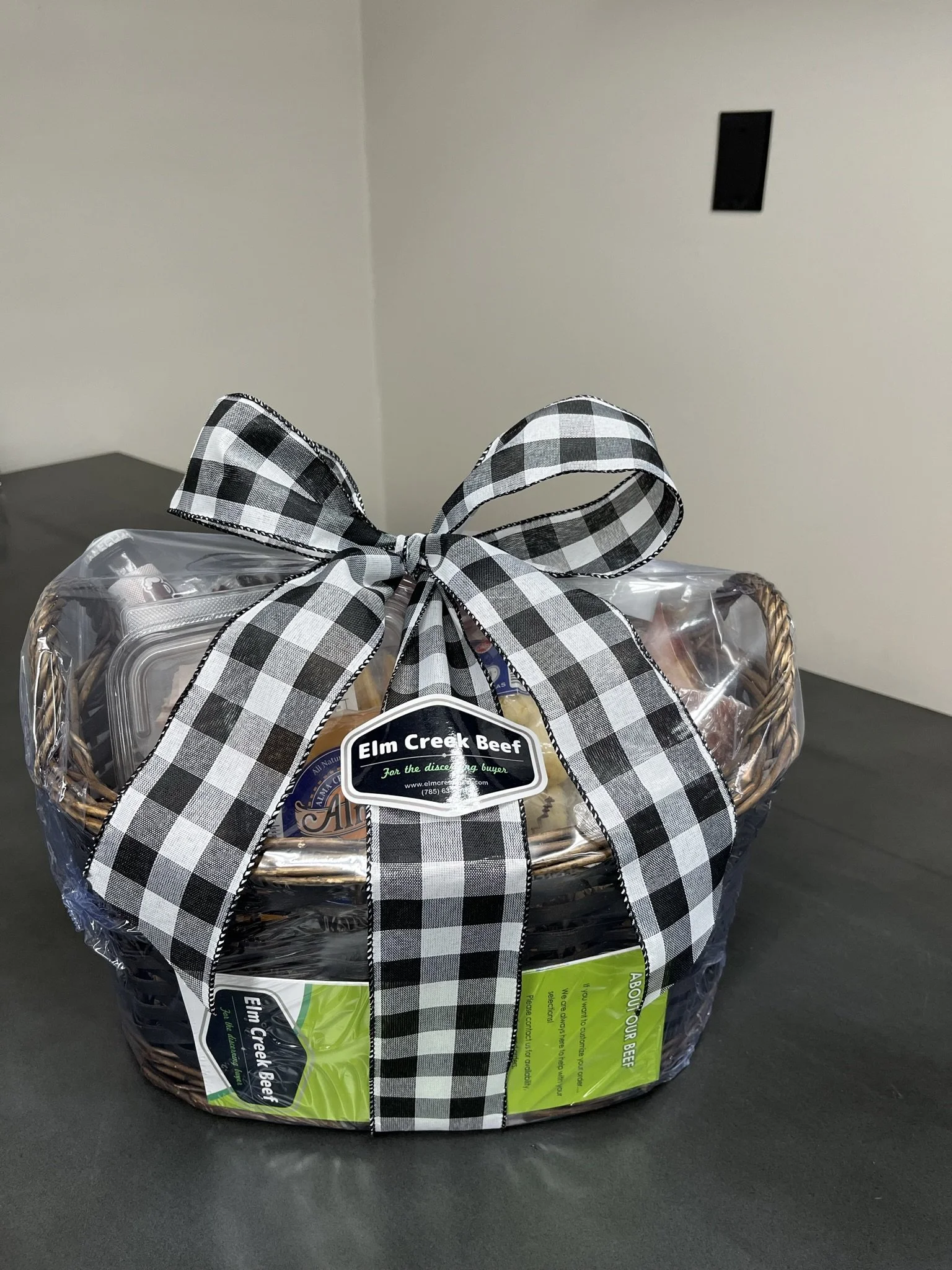 Elm Creek Beef A gift basket wrapped in clear plastic with a black and white checkered ribbon tied in a bow on top. The basket contains packages of Elm Creek Beef, with labels visible on the front.