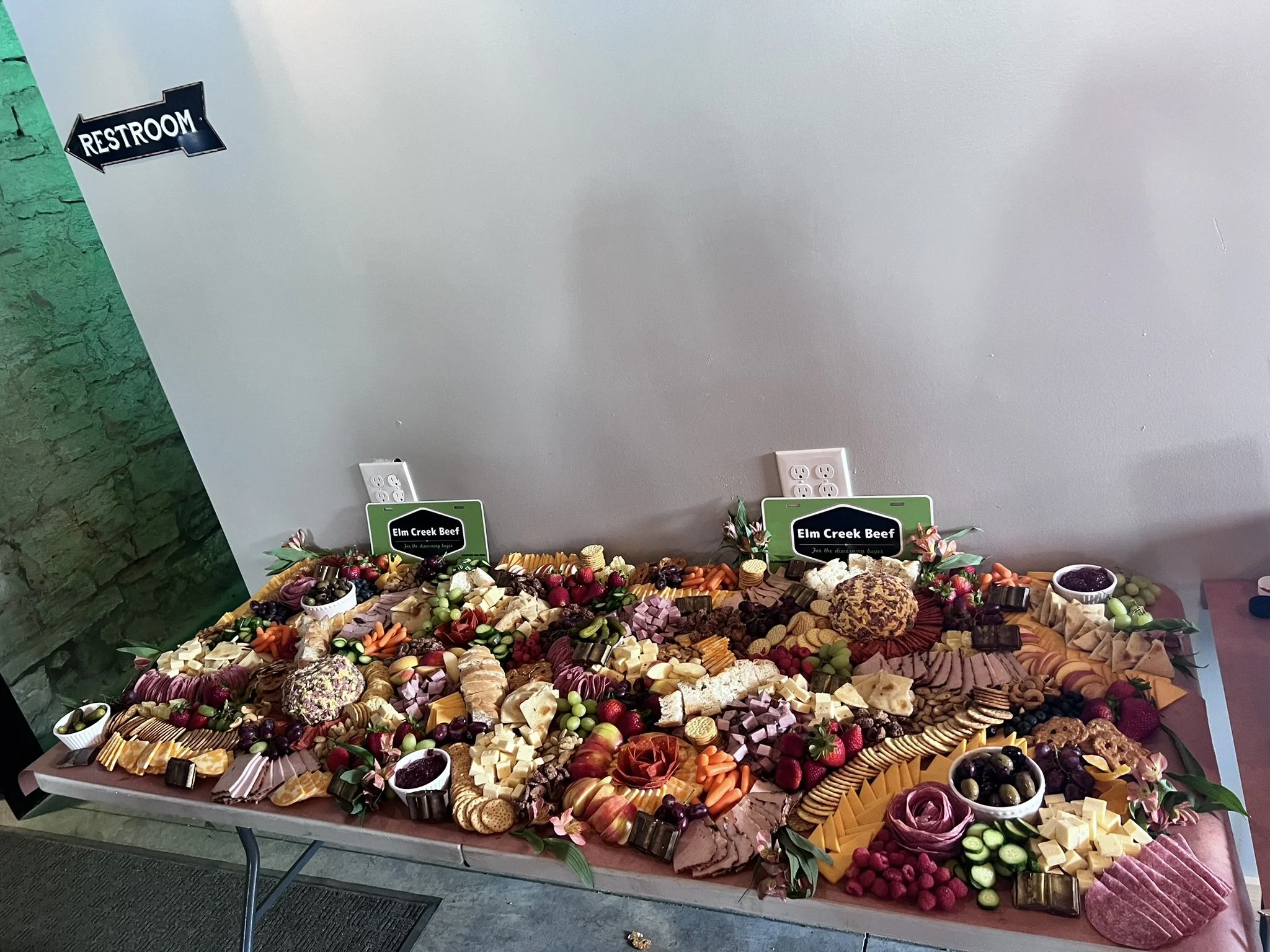 Elm Creek Beef Large tray of assorted cheeses, fruits, vegetables, crackers, and dips, with signs labeled 'Elm Creek Beef,' set on a table for a catering event.