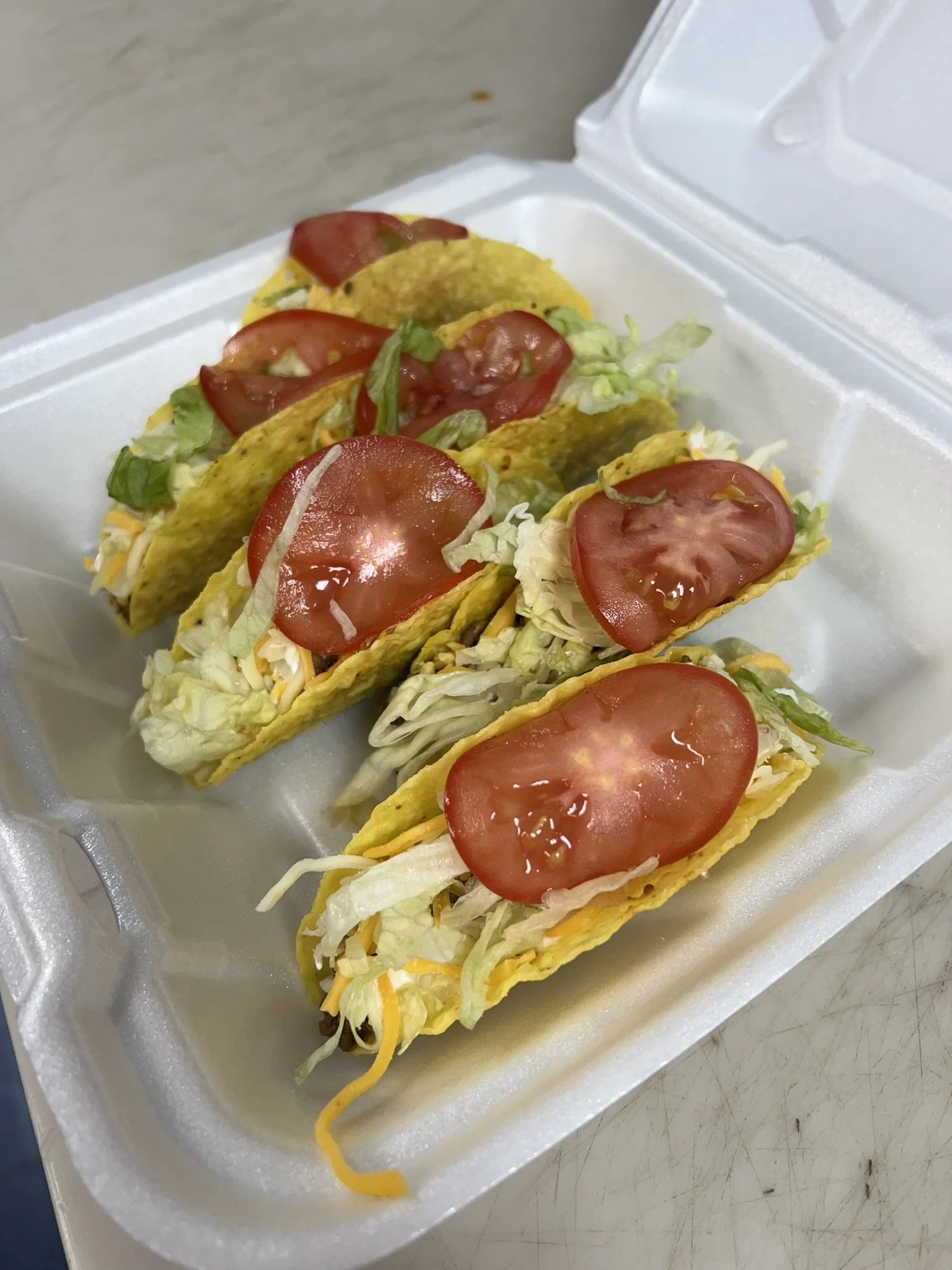 Elm Creek Beef Four tacos filled with lettuce, shredded cheese, and sliced tomato in a foam takeout container.