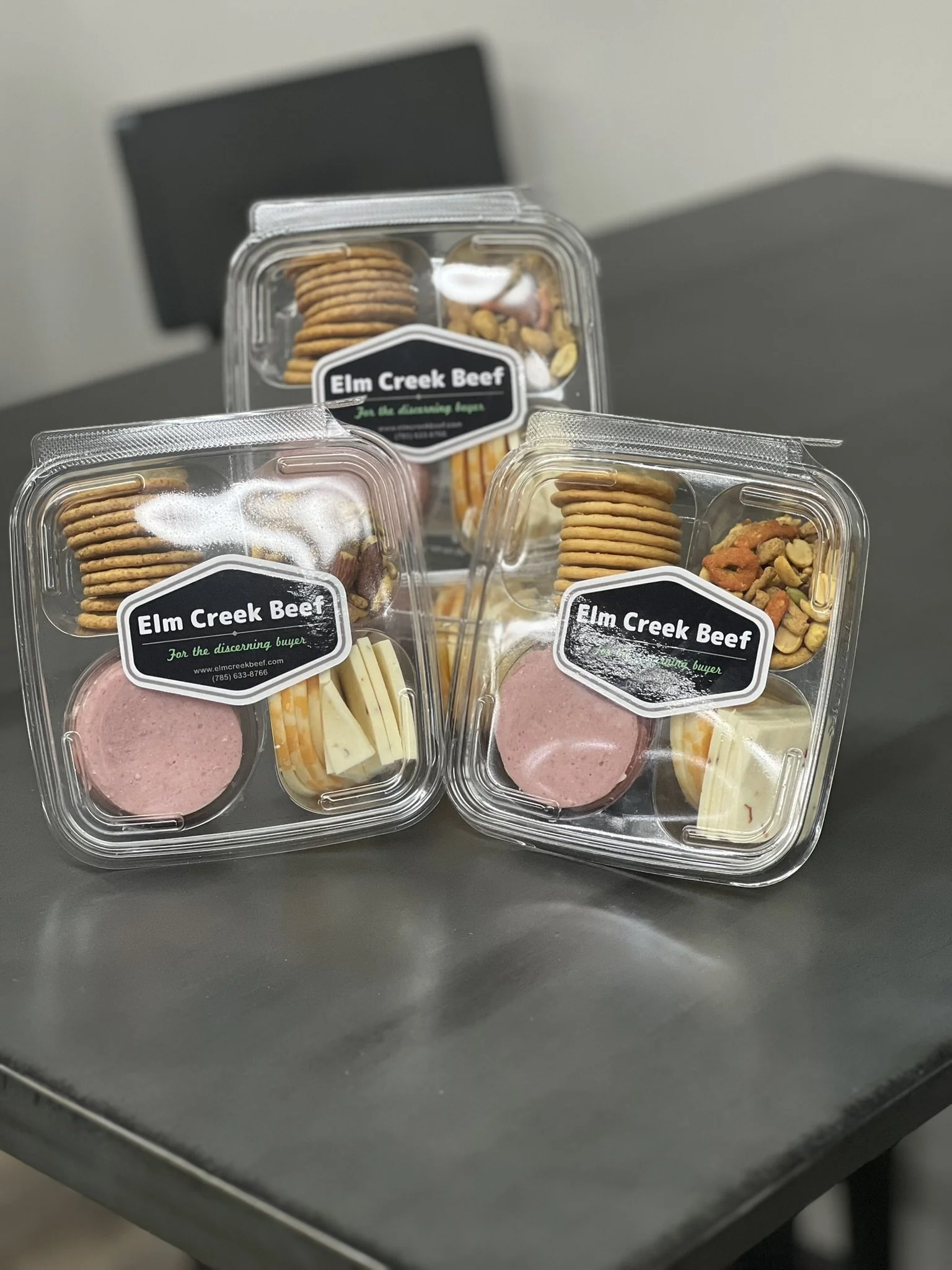 Elm Creek Beef Three plastic containers with assorted snacks and cheese, labeled 'Elm Creek Beef' on a table.