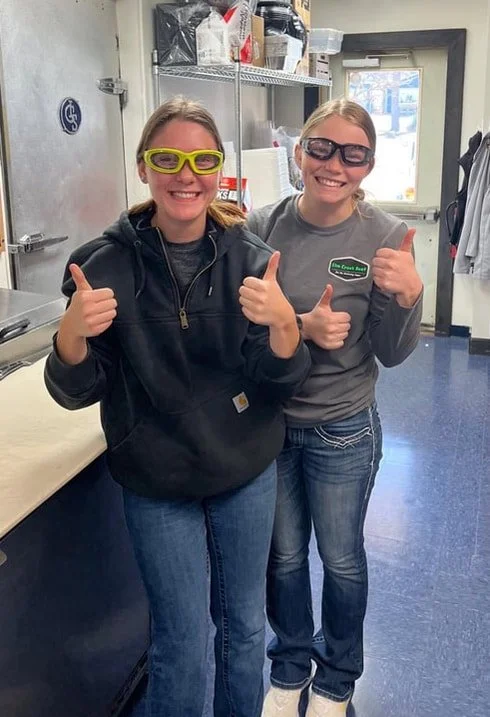 Two women standing inside a room, both wearing glasses and jeans, smiling and giving thumbs up. The woman on the left is wearing yellow glasses and a black hoodie; the woman on the right is wearing gray glasses and a gray shirt with a name tag.