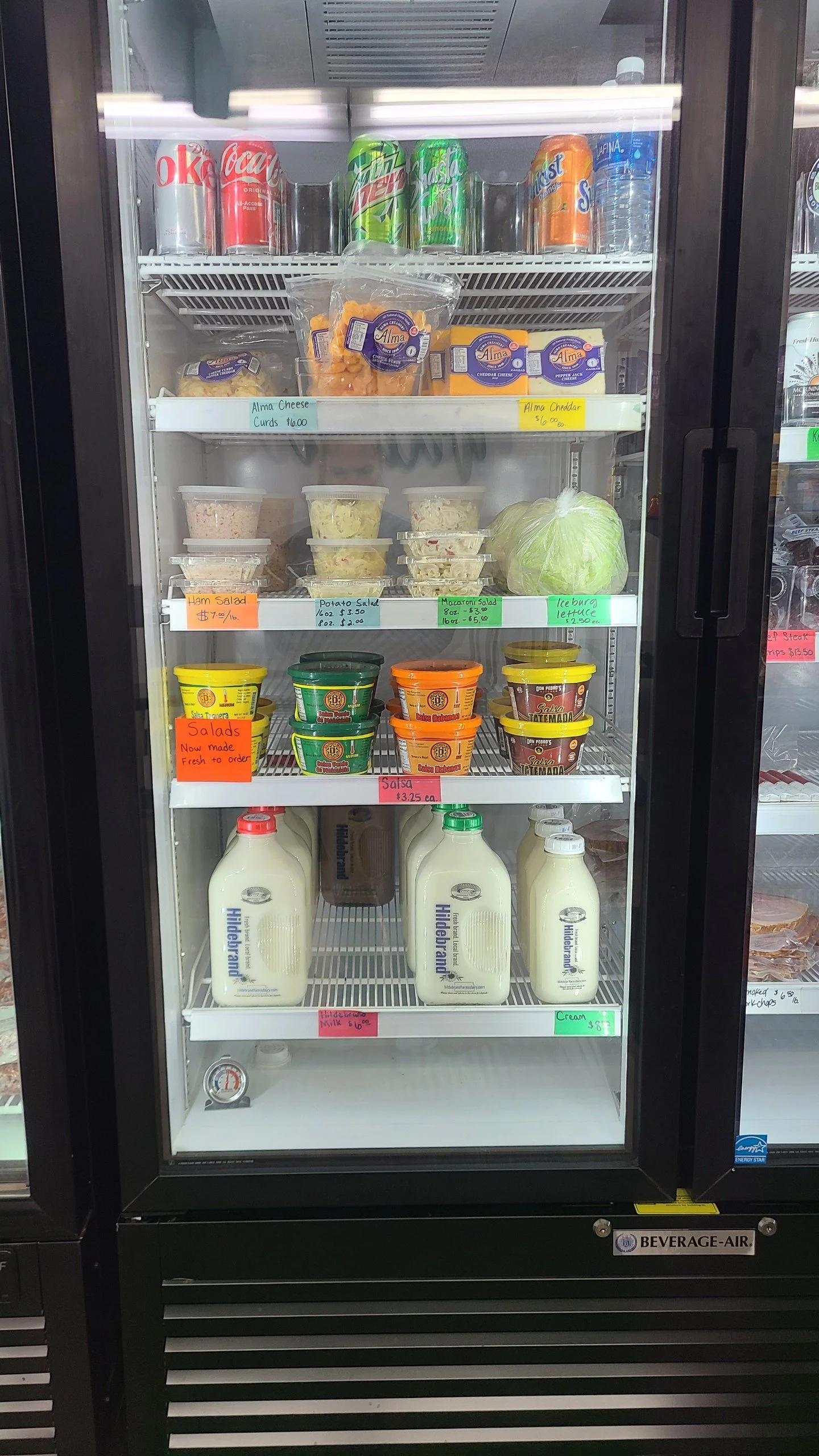 Elm Creek Beef A refrigerated display case containing various food and beverage items, including soda cans, bottles of milk, containers of salads, cheese, and sliced meats, with colorful handwritten price tags.