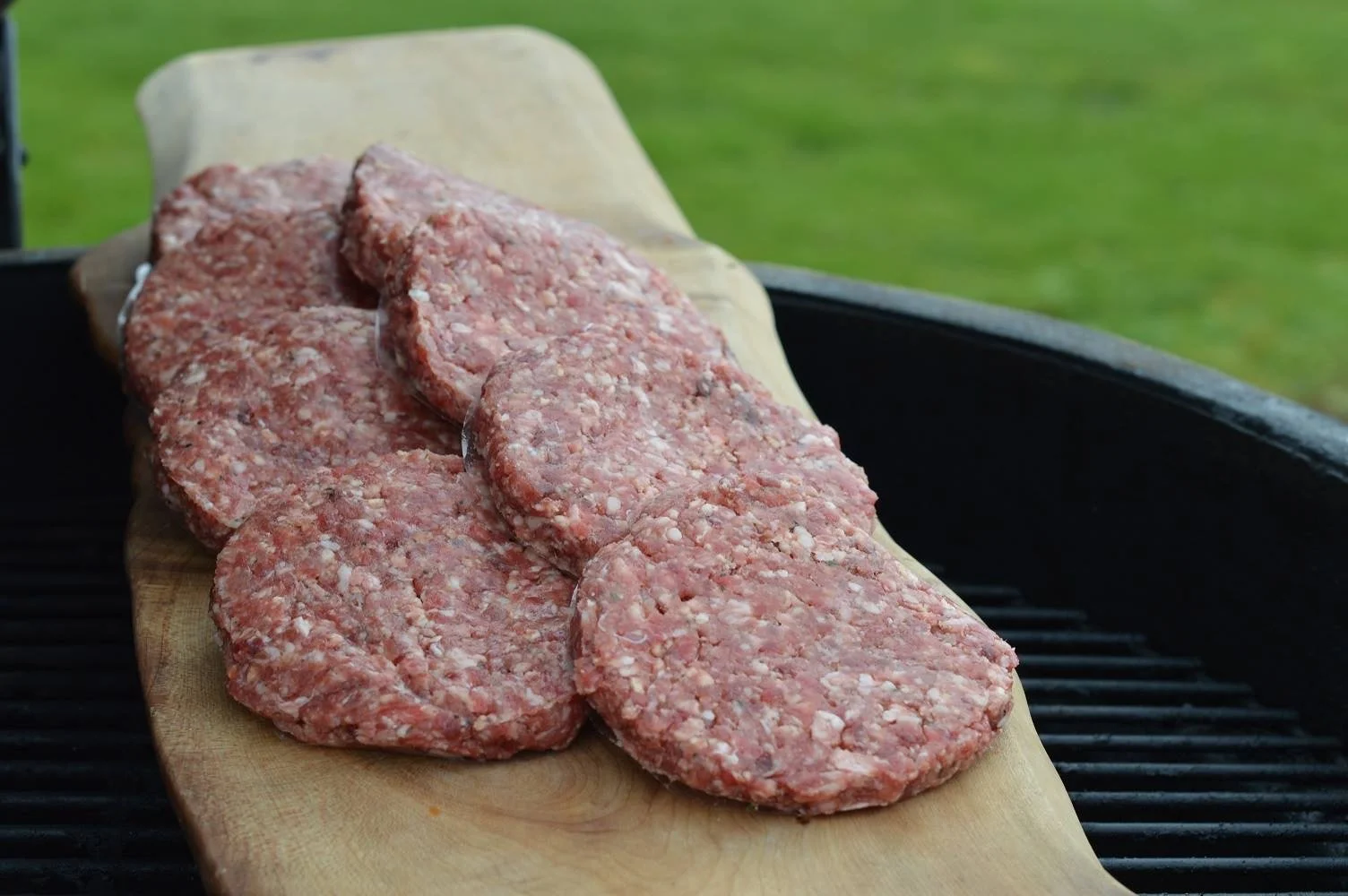 Elm Creek Beef Raw ground beef patties on a wooden board on a barbecue grill, with a green grassy background.