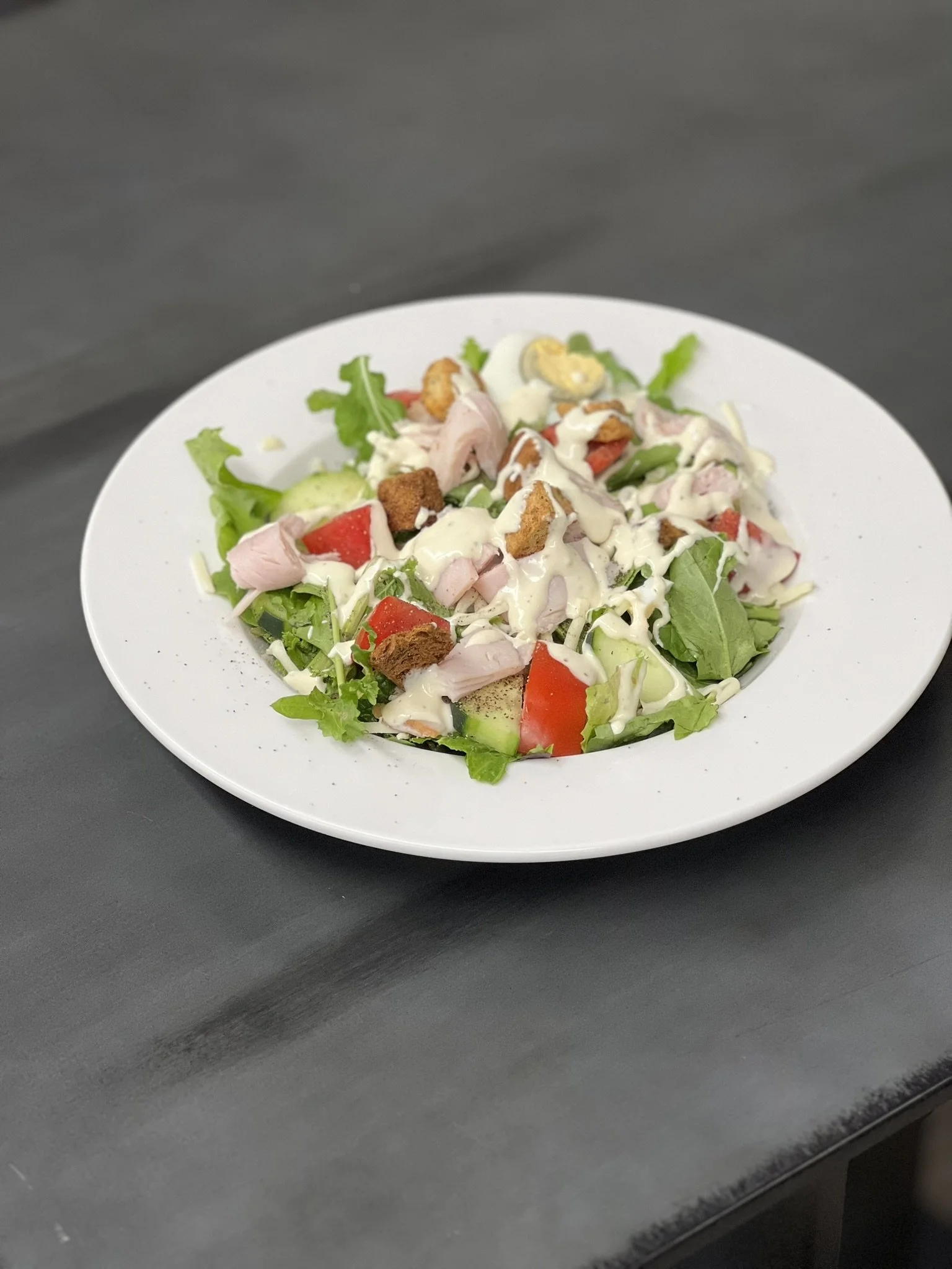 Elm Creek Beef A plate of tossed salad with diced tomatoes, cucumbers, lettuce, sliced chicken, hard-boiled eggs, croutons, and creamy dressing.