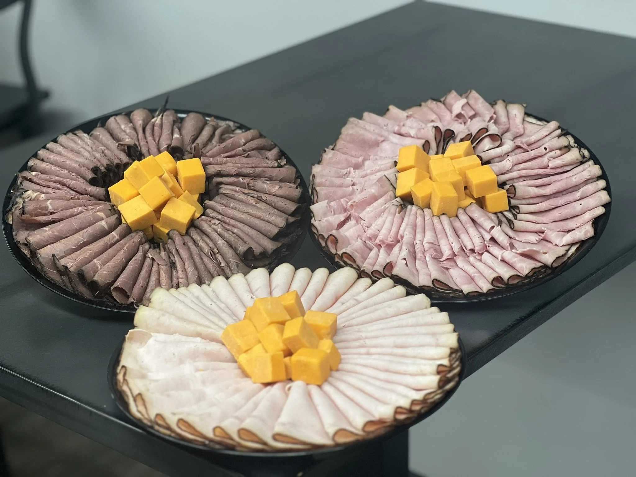 Elm Creek Beef Three black platters of assorted sliced deli meats topped with small cubes of cheddar cheese on a dark surface.
