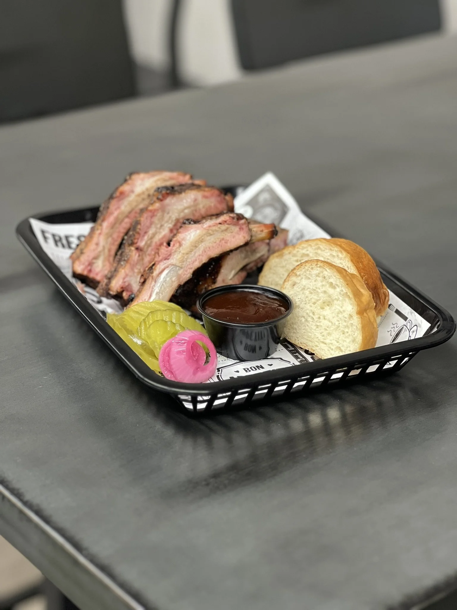 Elm Creek Beef A black tray with barbecue pork ribs, sliced bread, pickles, pink pickled onions, a cup of barbecue sauce, and a small container of baked beans, placed on a gray table.